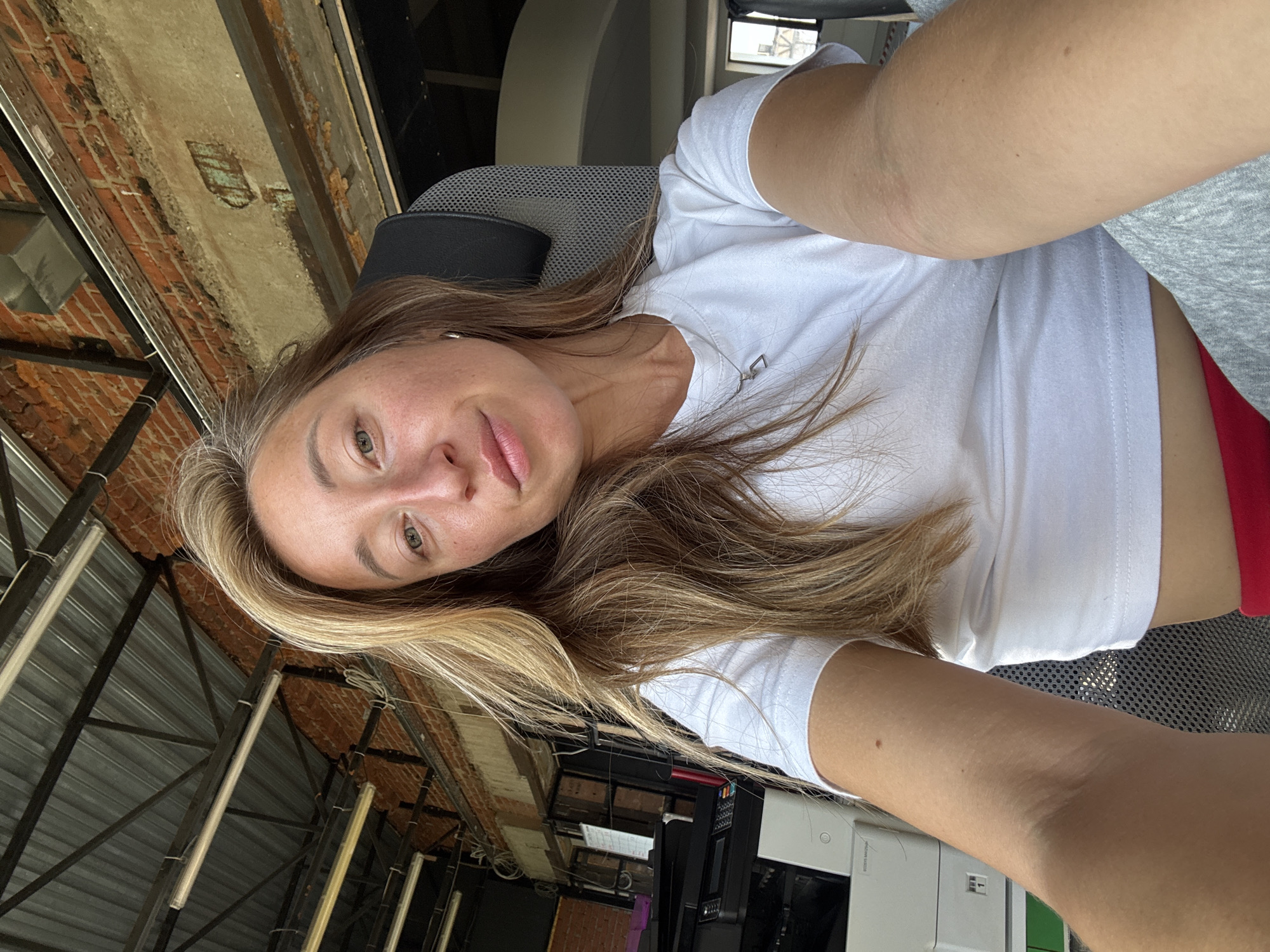 A selfie of a woman in Chaikovsky, Russia, shows her lying down in what appears to be an office or industrial setting. She has blonde hair and is wearing a white t-shirt. The background includes exposed brick walls, metal beams, and fluorescent lighting, suggesting an unfinished or open-plan space. The woman is looking at the camera with a relaxed expression. The lighting indicates it is daytime. No visible text is present in the image.
