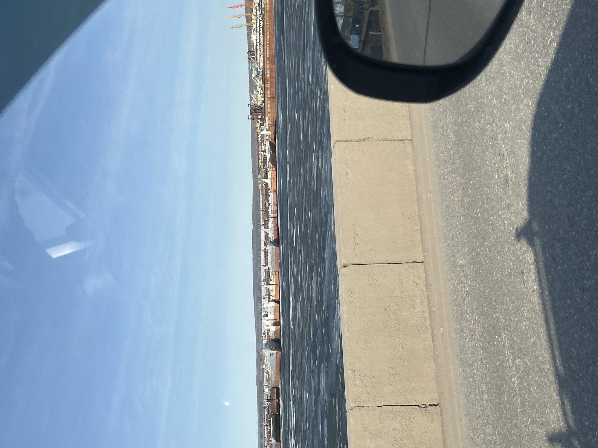 A sideways, eye-level shot taken from inside a moving vehicle, likely a car, shows a view of a canal or wide river with docks and what appears to be a large industrial area stretching along the opposite bank. The sky is clear and blue. A portion of the car's side view mirror and a section of the car's window frame are visible. The canal appears to have some ice or slush on the surface. Along the near side of the road, a concrete barrier separates the road from the canal. The photo was most likely taken in the city of Chaikovsky, Russia.