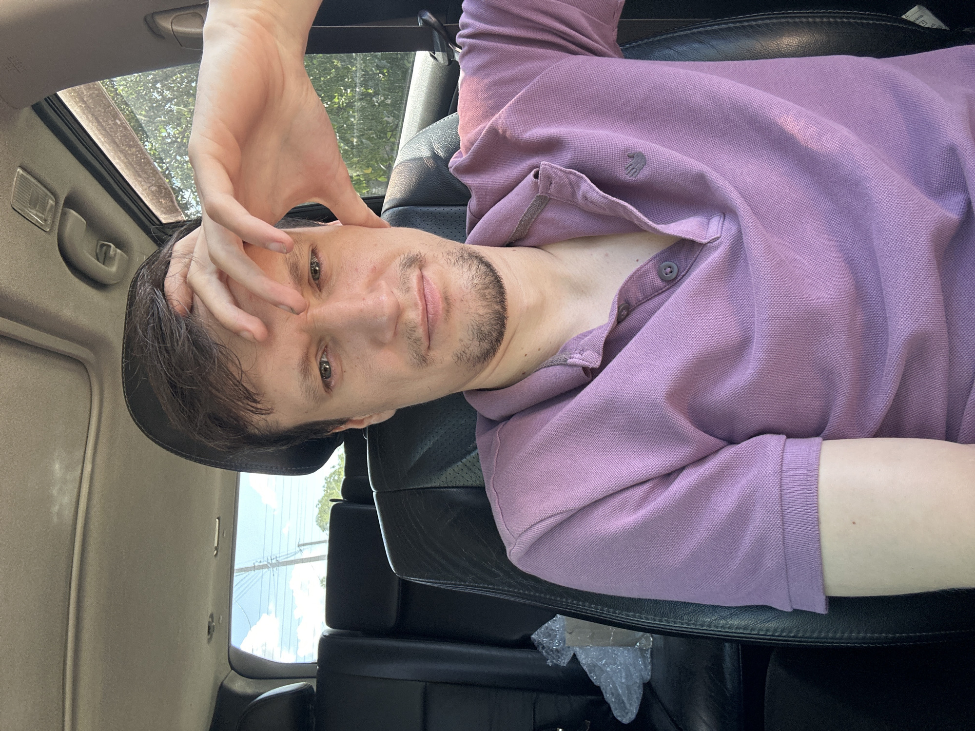 A man with light skin, dark hair, and a goatee is sitting in the driver's seat of a car. He is wearing a purple polo shirt and has his right hand on his forehead. His left arm is resting on his lap. The interior of the car appears to be black leather. Outside the car, there is a blue sky with white clouds, power lines, and trees. The scene is lit by natural sunlight. The image was taken in Makiv, Ukraine.