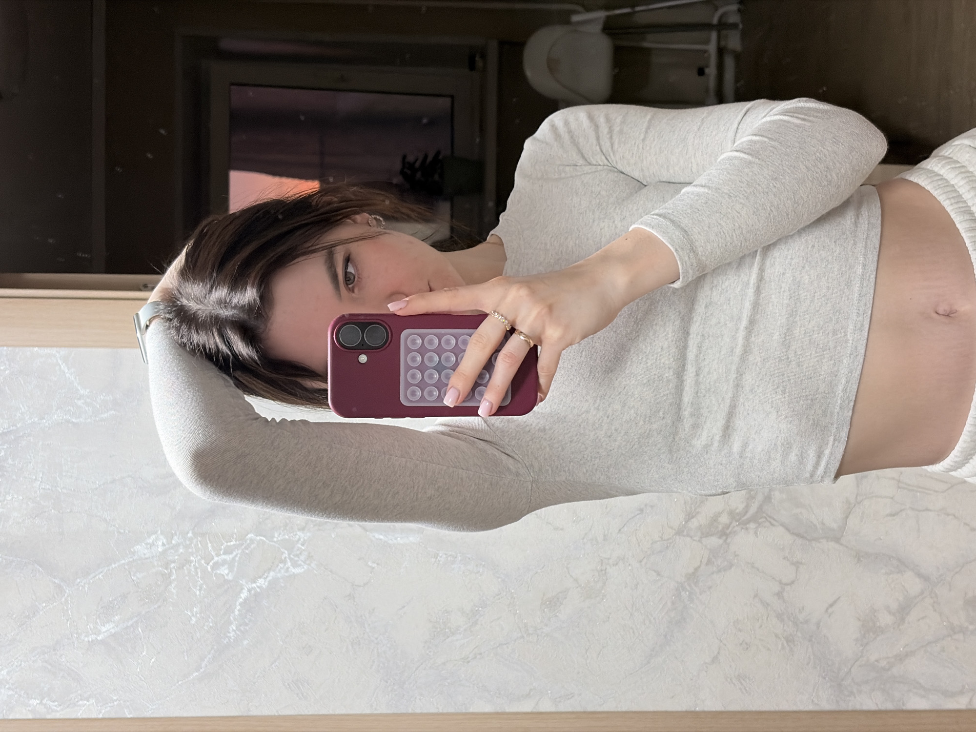 An indoor mirror selfie captures a young woman, partially visible, looking directly into the camera with a neutral expression. She has medium-dark brown hair and fair skin. She wears a light grey, long-sleeved crop top, revealing her midriff where faint stretch marks are visible around her navel. On her left wrist, she wears a light-colored watch or band. Her right hand, adorned with multiple rings, holds a dark red smartphone with a distinctive rectangular accessory featuring circular suction cups on its back. Her fingernails have a light pink or nude polish.
The reflection shows an interior space. Behind her upper body, a dark wall or furniture is visible, with a reflection of what appears to be a window or opening showing a reddish-orange glow, suggesting either sunrise or sunset. To her right, a light-colored object, possibly a toilet or bathroom fixture, is partially seen. The lower part of the scene consists of a white or light grey surface with a marble-like pattern, likely a countertop or wall, bordered by a thin, light brown wooden strip at the bottom of the frame. The overall setting appears to be a domestic bathroom or utility area. No visible text is present in the image.