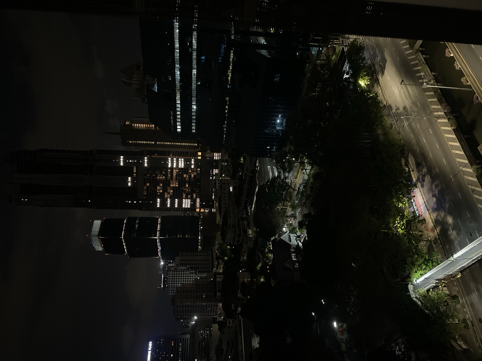 The image shows a nighttime view of a city skyline, specifically Bangkok, Thailand, as seen from a high vantage point. The cityscape is dominated by several tall buildings and skyscrapers with illuminated windows. The night sky is dark with clouds.
Below the buildings, there is a street with white pavement and lamp posts. Trees partially obscure the view of the street. A pedestrian bridge crosses the street.
The image appears to be taken from indoors, possibly from a window or balcony.
