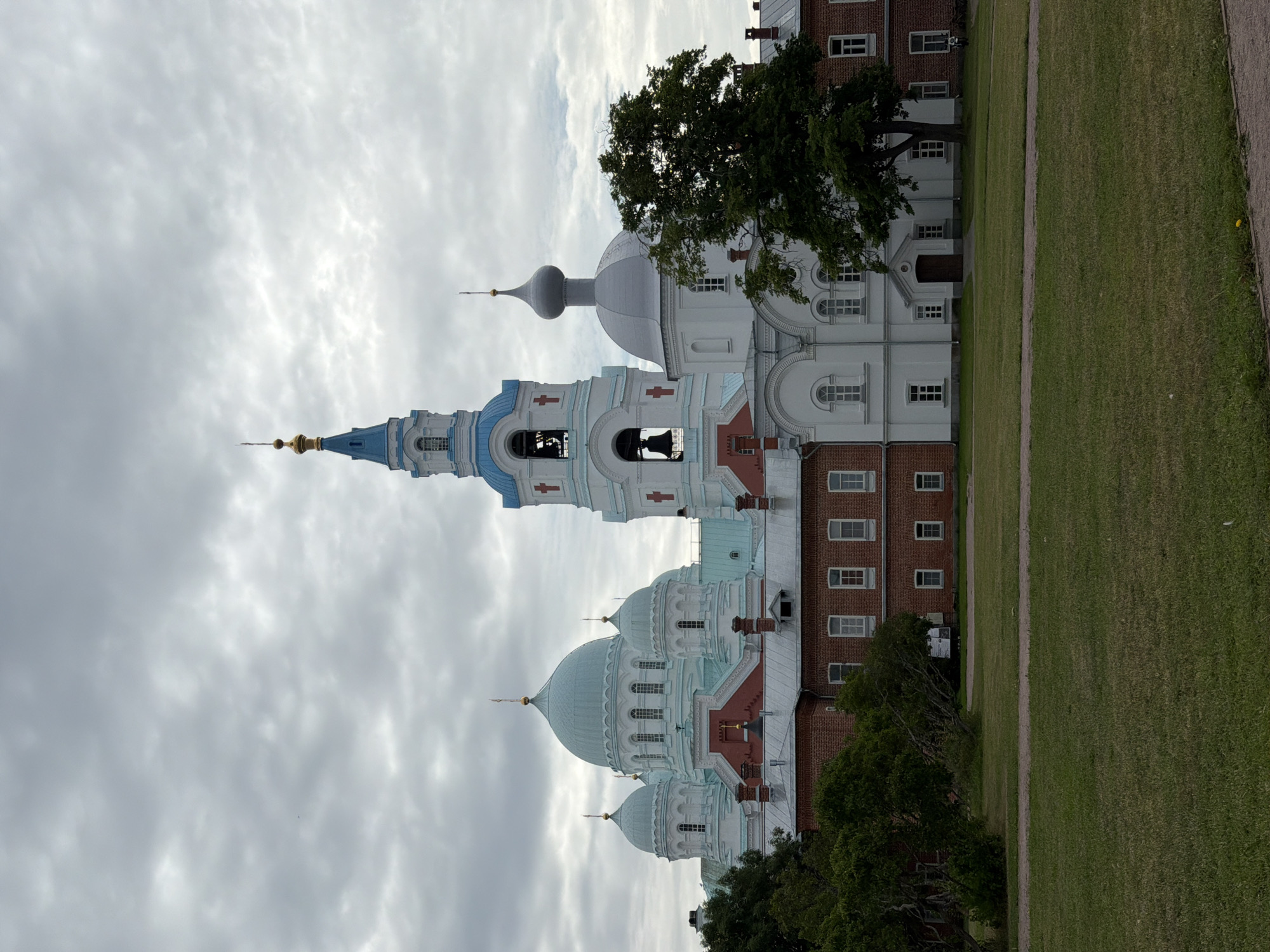 The image depicts a grand religious complex with multiple domes and a tall bell tower. The architecture features a blend of white and light blue structures with red brick buildings at the base. The main structures are adorned with golden crosses and intricate detailing. In the foreground, a well-maintained grassy lawn is visible, with several trees scattered throughout the scene. The sky is overcast with gray clouds. The image is taken from a slightly low angle, emphasizing the scale of the buildings.