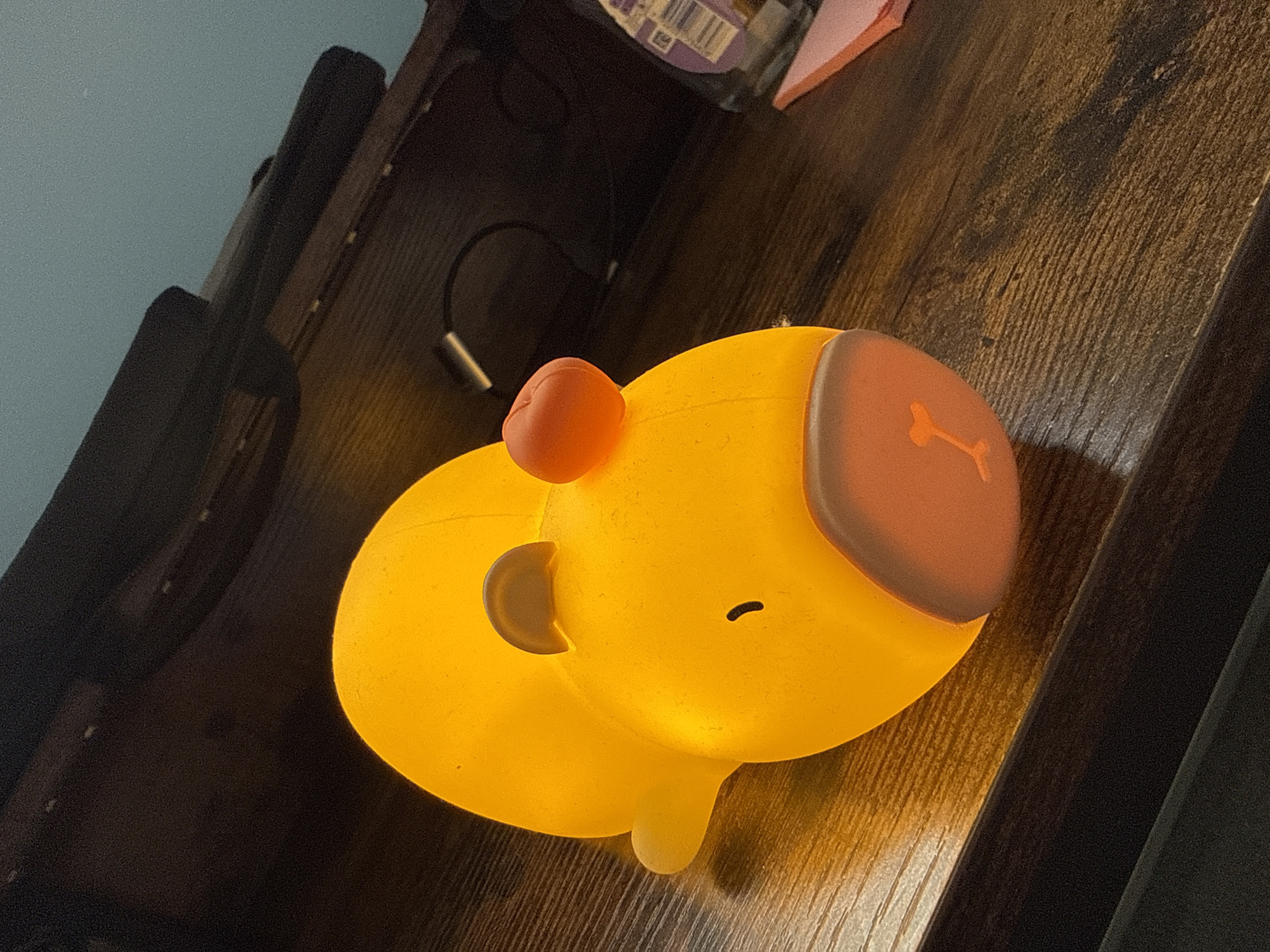 A yellow night light shaped like a capybara is on a brown wooden surface. The capybara has a red nose, a bone design on its head, and a small tail. A black charging cord is visible in the background. The image suggests an indoor setting, possibly a bedroom or a study. The location appears to be inside a building, but no further location specifics are available.