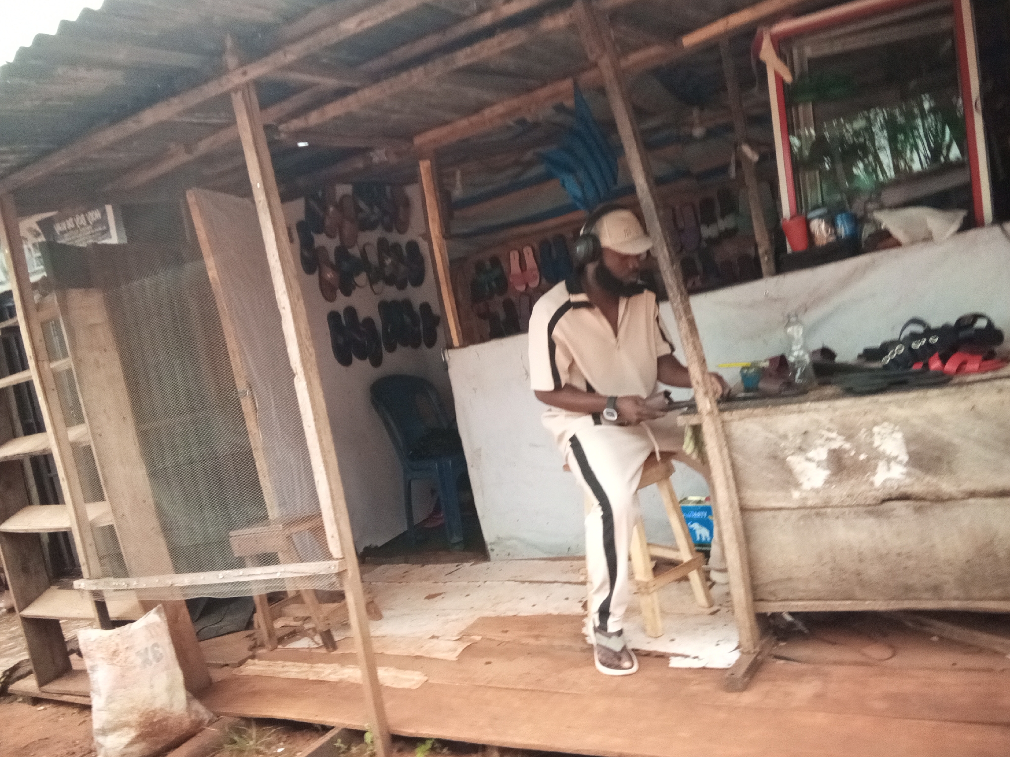 The image depicts a man wearing a light-colored shirt and pants, a cap, and headphones, standing in what appears to be an outdoor market stall in Oko Town, Nigeria. He is holding a device, possibly a phone, in his right hand and looking down at it. The stall is constructed from wood and corrugated metal, with shelves displaying various items, including what seem to be shoes and sandals. A blue chair is visible in the background, and there are bags and other objects scattered around the area. The ground is made of rough wooden planks and dirt. The overall impression is of a functional, possibly informal, retail space in a developing urban environment.