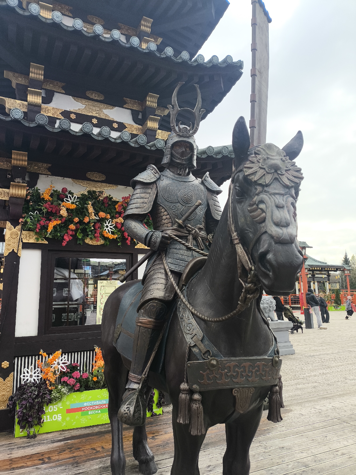 A large, dark bronze statue of a samurai warrior on horseback stands prominently in the foreground, facing slightly right. The samurai is depicted in full, detailed armor, including an ornate helmet with prominent crests, a breastplate, and arm guards. Two swords are visible at his left hip. The horse also wears decorative armor, featuring an intricate headpiece, an ornate saddle, and detailed chest and flank coverings with tassels.
Behind the statue is a multi-tiered building showcasing traditional Japanese architecture, characterized by dark wooden beams, curved eaves, and ornate roof tiles with circular crests. Gold and white decorative elements adorn the fascias. A window below is partly obscured by a planter box filled with vibrant artificial flowers in shades of orange, red, and purple, interspersed with white star-shaped cutouts.
On this green planter box, Cyrillic text is visible, advertising "ФЕСТИВАЛЬ МОСКОВСКАЯ ВЕСНА" (Moscow Spring Festival) with dates "01.05 - 11.05".
In the background, under an overcast sky, more people can be seen strolling on a wooden boardwalk or paved area, and further Japanese-style buildings with red torii-gate-like structures are faintly visible. A tall, light-colored banner or flag pole stands to the right of the main building. A small, white sculpted lion-like figure rests near the horse's front leg. The scene takes place outdoors during the daytime under cloudy conditions, in what appears to be a Japanese cultural garden or exhibition space in Moscow, Russia.