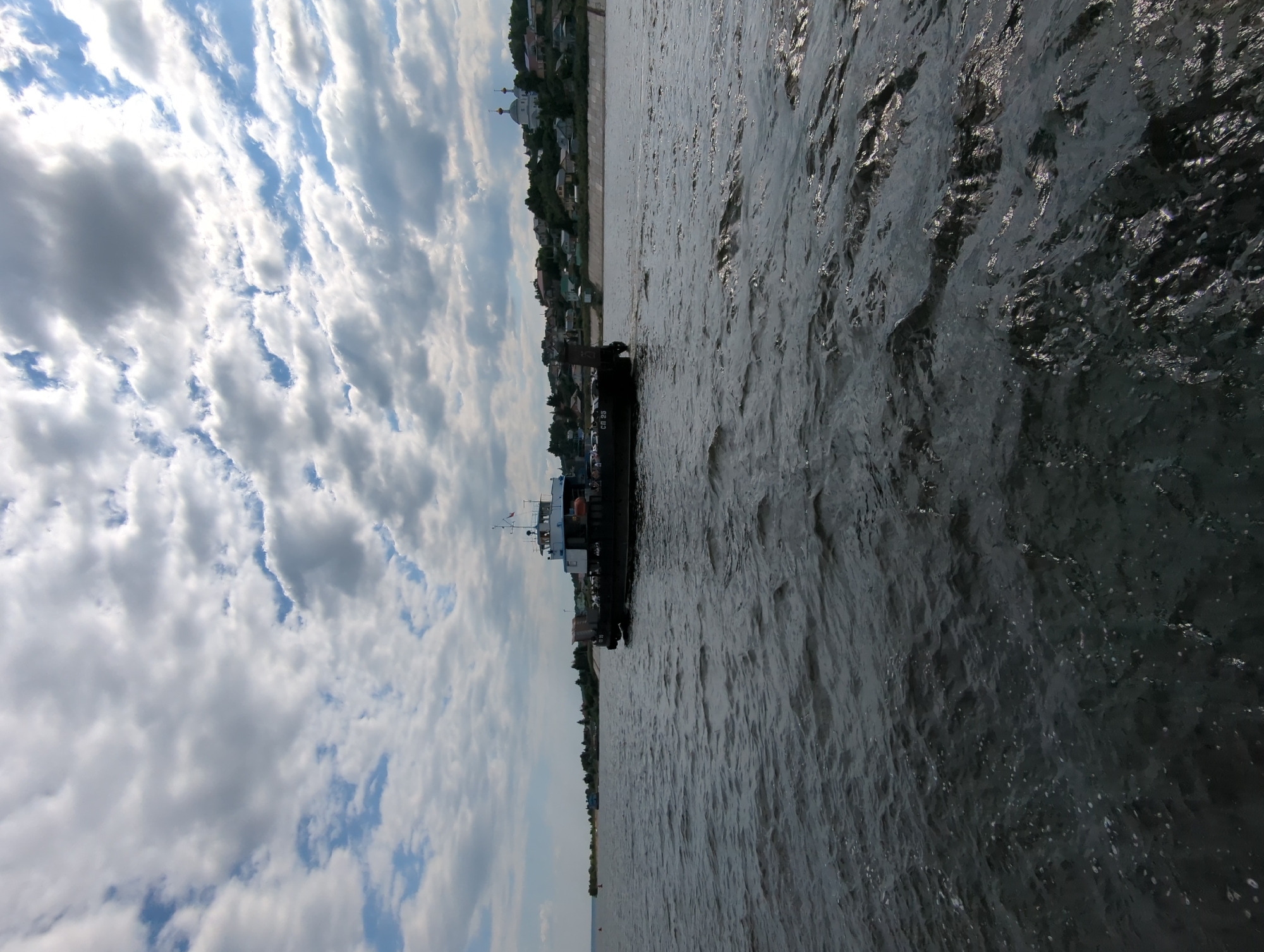 The image shows a ferryboat on a body of water, likely a river or lake, with a town or village on the distant shore. The sky is filled with dramatic, textured clouds, some dark and some bright white, suggesting a dynamic weather pattern. The water is choppy, with ripples and reflections of the sky and the ferry.
The ferryboat is dark and appears to be carrying vehicles or cargo, with several people visible on its deck. It is positioned in the middle ground of the image, moving from left to right. The shoreline on the right side of the image features a collection of buildings, including houses and what appears to be a church with domes, nestled on a hillside. Trees are also visible along the shore. The far left of the image shows a less developed shoreline with some scattered structures. The overall impression is of a scenic, possibly rural or semi-rural, setting.
