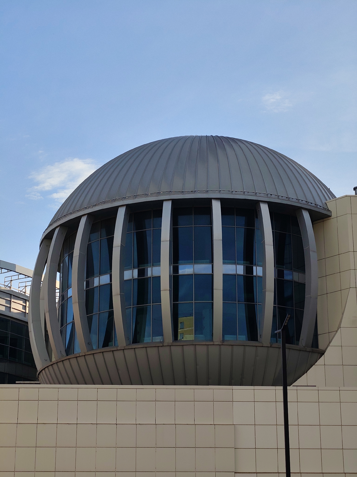 This image captures a striking modern architectural structure in Moscow, Russia, dominated by a large, metallic dome. The main subject is a building featuring a prominent, grey, ribbed dome at its summit, reflecting the clear blue sky.
Below the dome, there is a distinct curved section characterized by tall, vertical, and slightly bowed metallic structural elements. These elements frame large, reflective glass windows that mirror the blue sky and faint hints of surrounding buildings, including a yellowish reflection in one pane. Further down, another grey, ribbed metallic band forms the base of this curved section. The lower visible part of the building's facade consists of numerous light-colored, possibly off-white or beige, square tiled panels, creating a textured surface. To the right of the main dome structure, another section of the building with similar light-colored paneling is visible, connecting to the central dome. Hints of other modern building components can be seen on the left side of the frame.
The photograph appears to be taken during the daytime under fair weather conditions, indicated by the bright illumination and the clear blue sky with only a few wispy clouds. The perspective is slightly upward, emphasizing the scale and grandeur of the dome. A slender black pole, possibly a lamppost or part of urban infrastructure, stands vertically in the lower right foreground, partially obscuring the building. No people or discernible activities are visible, and there is no visible text on the building or in the scene. The overall impression is one of modern, clean design and imposing scale.