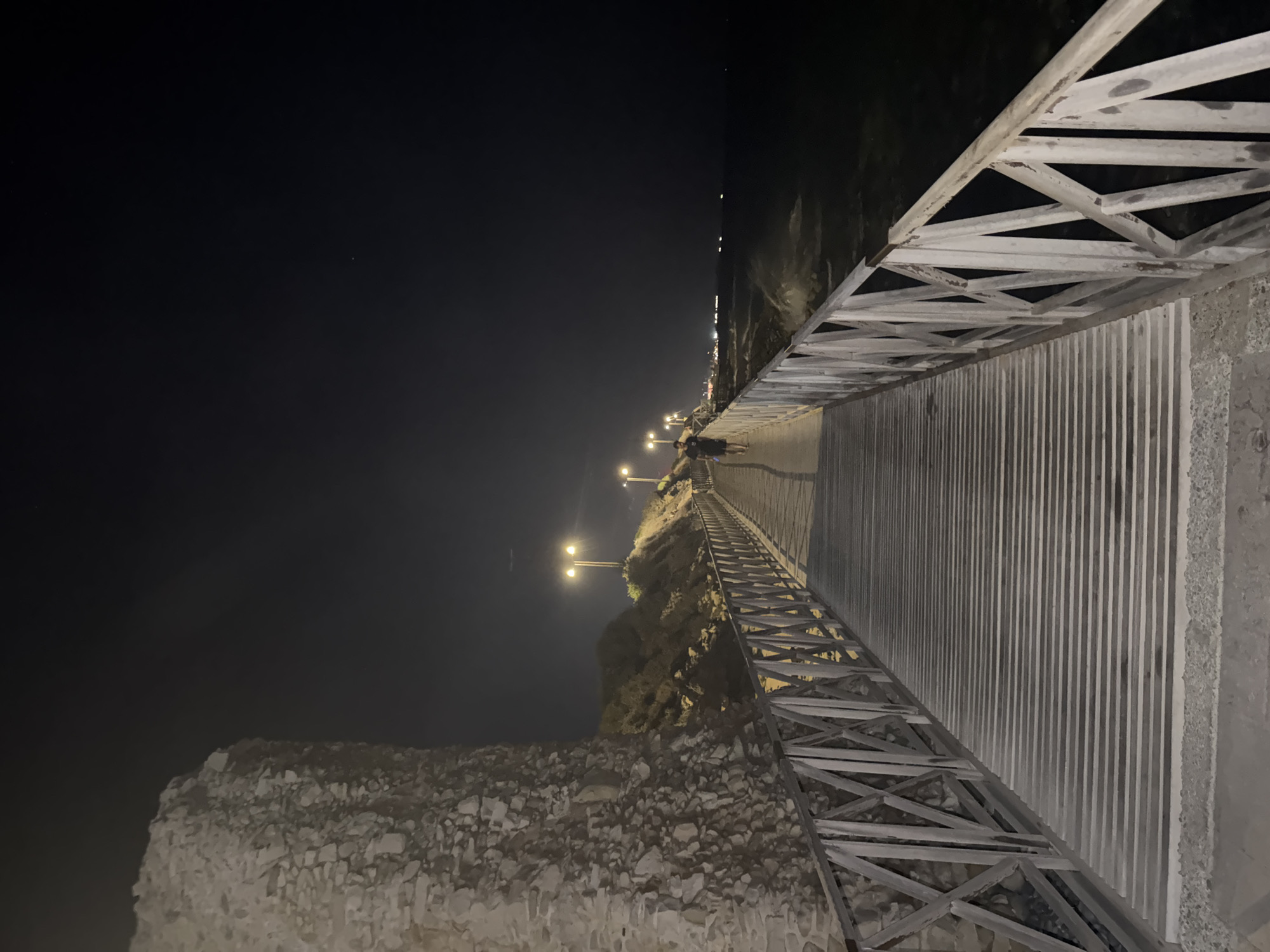 The image shows a boardwalk at night in Agios Tychona Community, Cyprus. A stone structure, possibly part of an ancient ruin or fortification, is visible on the left. A wooden boardwalk with a crisscross railing extends into the distance, illuminated by streetlights. A lone figure is walking on the boardwalk. The sea is to the right, with waves crashing against the shore. The sky is dark, with a few faint stars visible. The overall scene suggests a coastal walkway or promenade at night.