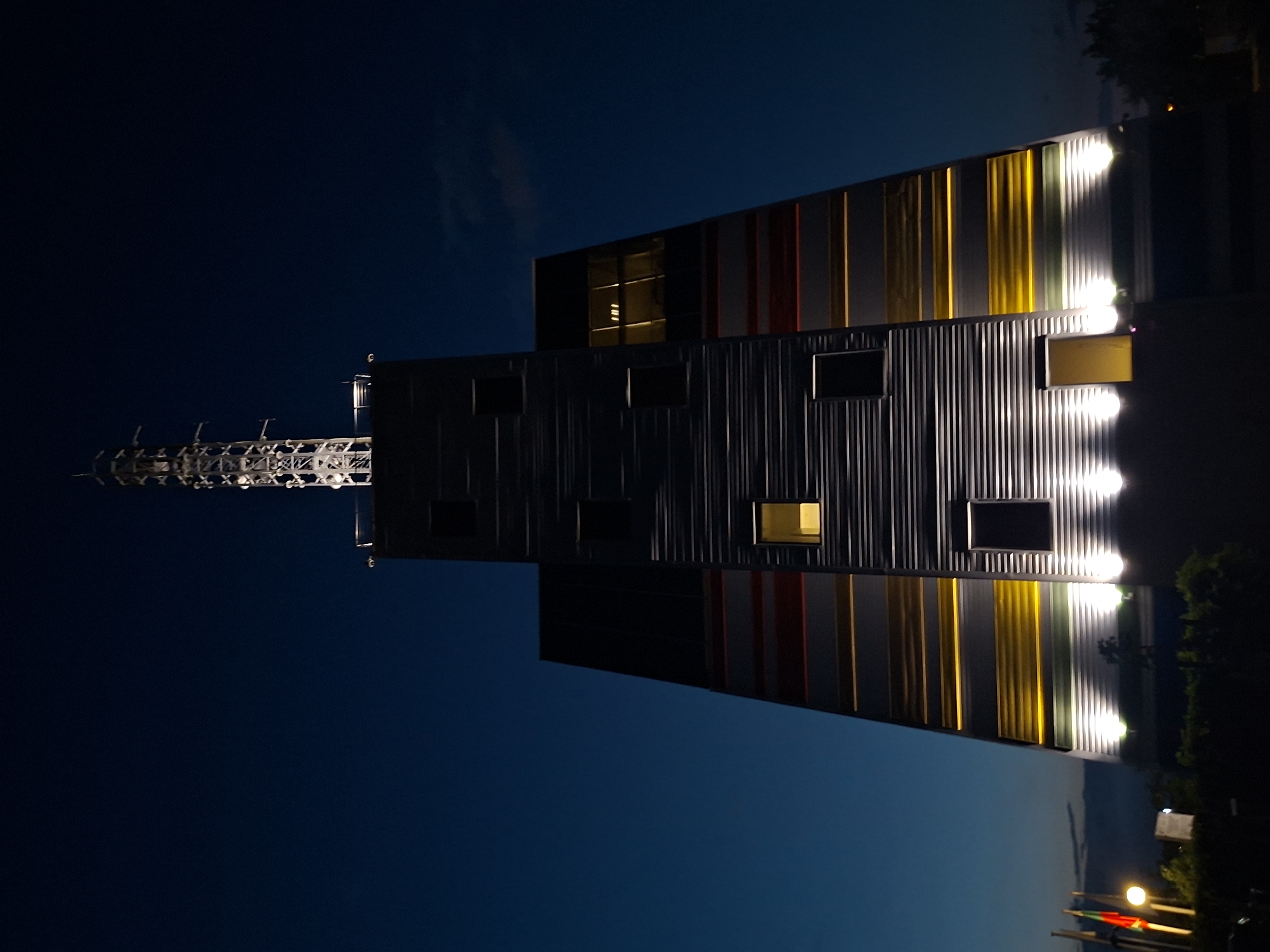 The image depicts the Radio Television Tower of Varna, Bulgaria, at night. The tower, a prominent landmark, is illuminated, showcasing its modern architecture. The lower portion of the tower features horizontal stripes of red, yellow, and white, set against a darker background. Several windows are visible, with one on the left side glowing with a warm yellow light. The base of the tower is lit by spotlights, casting light upwards onto the structure. A tall antenna mast rises from the top of the tower, silhouetted against the dark blue night sky. In the foreground, two flags are visible on poles, one appearing to be the flag of Bulgaria. Trees and bushes are present in the lower foreground, and the general environment suggests an outdoor setting at dusk or night. The scene is devoid of people, and no specific actions or interactions are captured.