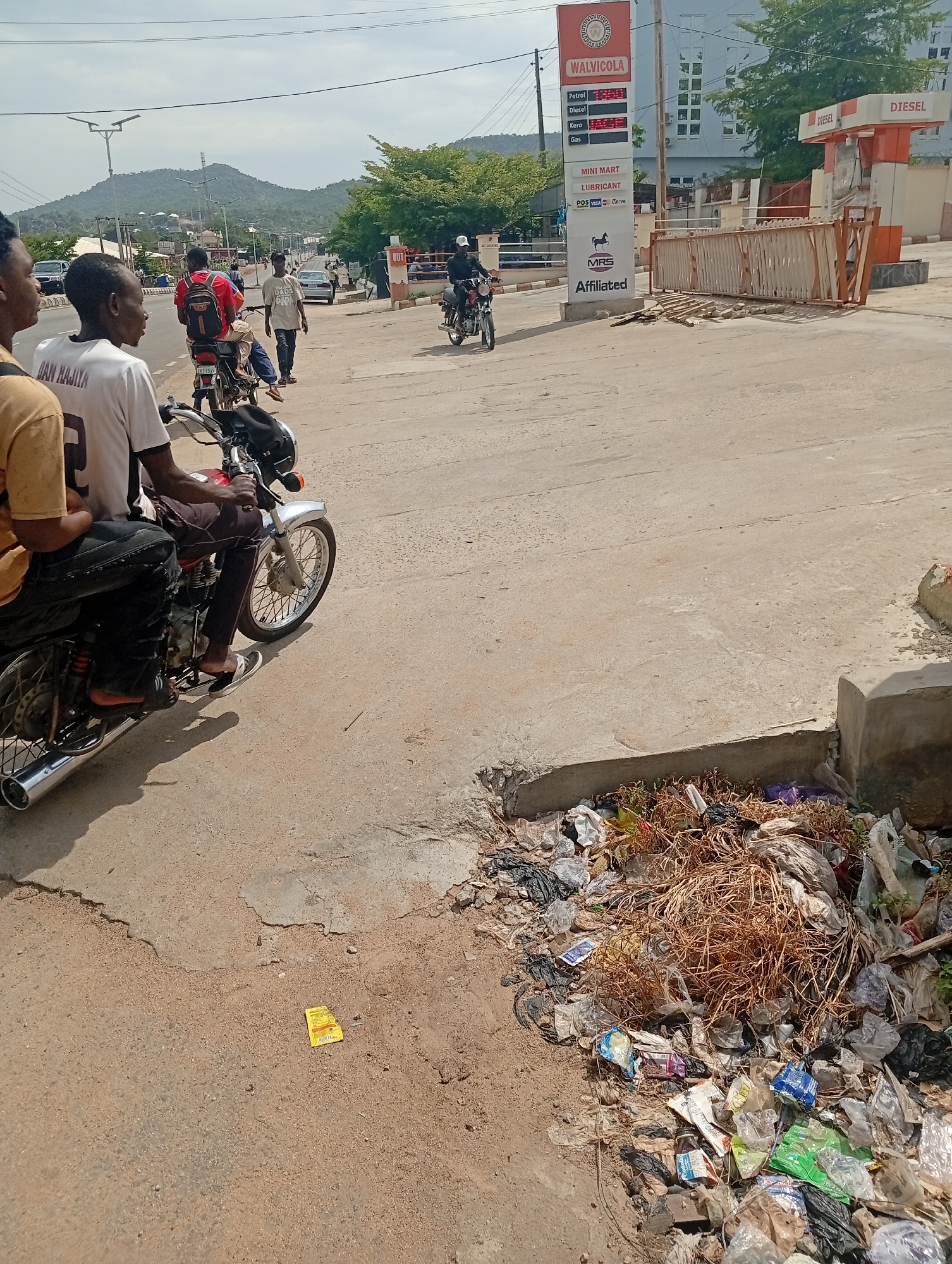 The scene depicts a street in Adankolo, Nigeria, with a view of a Walvicola petrol station. The street is concrete, and on the right side, there's a pile of mixed garbage, including plastic bags, paper, and dry vegetation. Several people are present: two are riding a motorcycle in the foreground, another rides one further down the street, and others are walking on the side of the road. A red sign labeled "DIESEL" is near the station. The petrol station sign displays prices for Petrol, Diesel, Kero, and Gas, and includes logos for Mini Mart, VISA, Verve, and MRS Affiliated. The sky is overcast, and in the distance are mountains.