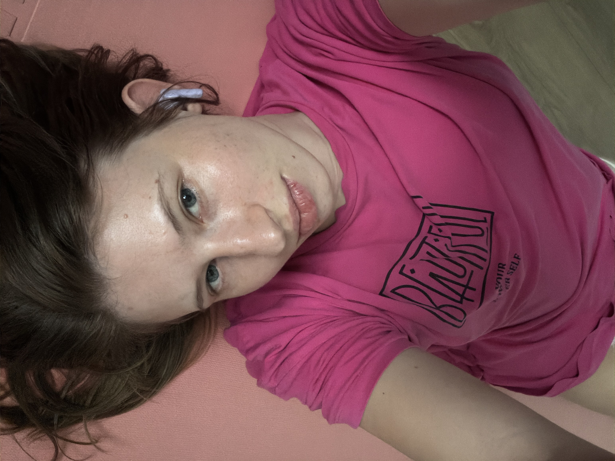 A person is lying on their back on a pink yoga mat. The person is wearing a bright pink t-shirt with a black graphic design on the front. The person's head is tilted to the side, and they are looking upwards. The person has fair skin, blue eyes, and brown hair. There is a slight sheen of sweat on the person's forehead and cheeks. The background consists of a wooden floor and a wall. The image appears to be a selfie, taken from a high angle. The specific location of Dongducheon-si, South Korea is not discernible from the image itself.