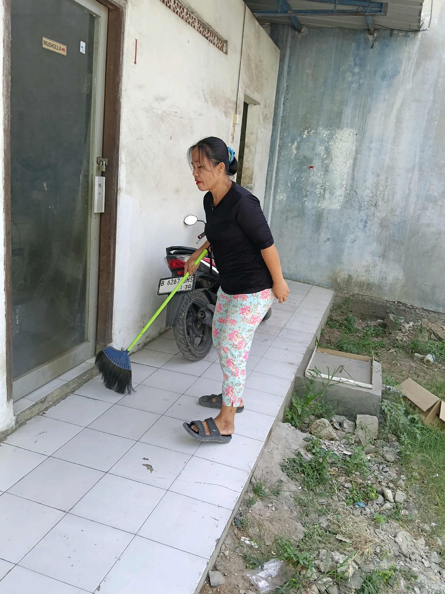 A medium shot captures a woman in a black t-shirt and floral-patterned leggings sweeping the tiled ground outside a building. She is facing left, leaning forward with a green-handled broom. To her left is a closed glass door with a sign reading "MUSHOLLA" and an arrow pointing right. Behind her, a motorcycle is parked. The building's wall, to the right, is painted in shades of blue and gray with peeling paint. The ground is a tiled pathway bordering a dirt area with sparse vegetation and debris, including a cardboard box and some rocks. The scene appears to be in Belimbing, Indonesia, given the presence of the motorcycle's license plate which starts with "B".