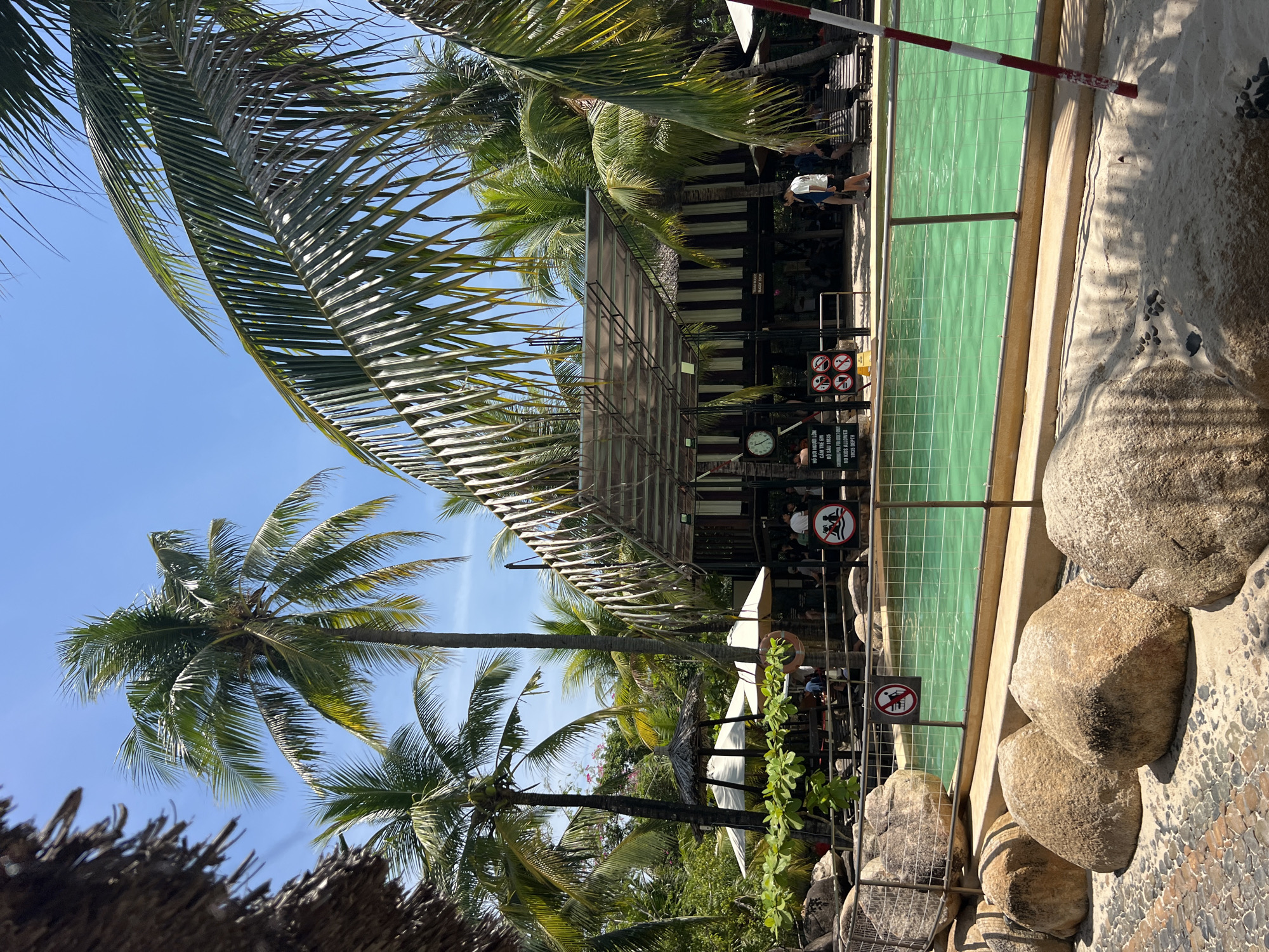 The image depicts a vibrant outdoor swimming pool area, likely a resort or recreational facility, set against a backdrop of lush tropical vegetation and clear blue skies. Tall palm trees with fronds rustling gently in the breeze dominate the scene, creating a distinct tropical ambiance. The pool itself is filled with bright, turquoise water, inviting and reflecting the sunlight.
In the foreground, large, weathered rocks are scattered across a sandy and pebbled ground, adding a naturalistic element to the landscape. A metal fence with a safety net separates the pool area from the foreground, with several circular signs indicating pool rules. One sign, partially visible, shows a silhouette of a person being cautioned. Another sign displays a clock and text in Vietnamese, potentially related to operating hours or pool depth regulations.
Behind the pool, a covered structure with a transparent roof and dark wooden accents serves as a shaded area, with people visible underneath, suggesting a dining or relaxation space. Lounge chairs are arranged nearby, indicating a place for sunbathing. Several individuals, dressed casually, are present in the background, some walking and others appearing to be seated. The overall impression is one of a lively and well-maintained leisure destination, perfect for a sunny day. The mention of Tay Nha Trang Ward, Vietnam, places this scene in a coastal city known for its beaches and resorts.