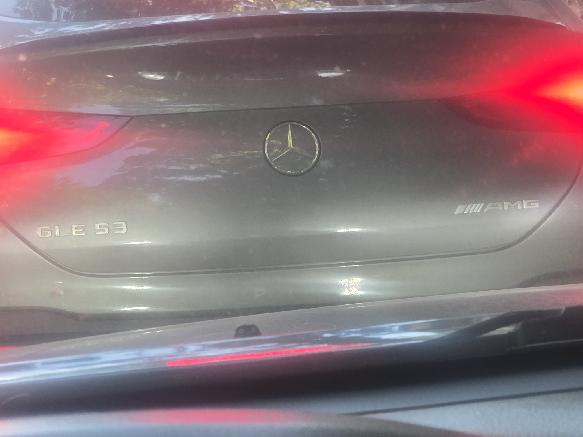 The image shows the rear of a gray Mercedes-Benz GLE 53 AMG. The Mercedes-Benz logo is visible in the center. The "GLE 53" badge is located on the left side, and the "AMG" badge is located on the right side. The brake lights are illuminated. The photo was taken from inside a vehicle, looking out through the windshield. The location is Unknown, Unknown.