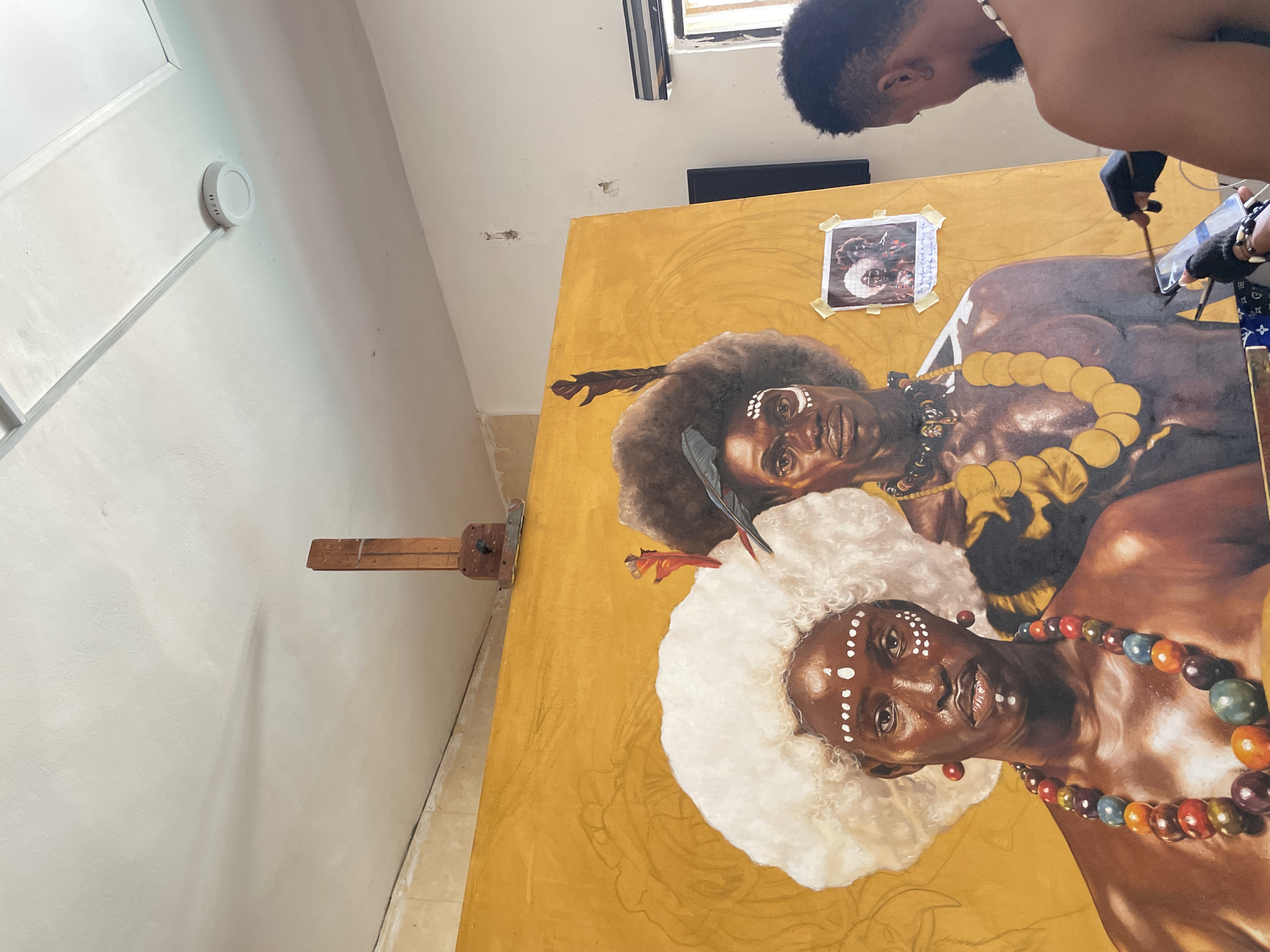 The image captures a scene inside an art studio in Ado, Nigeria. In the foreground, a large canvas depicting two dark-skinned figures wearing traditional attire is in progress. The figures are adorned with beads, feathers, and white facial markings. A partially visible artist is working on the upper section of the canvas, using a paintbrush and a reference photo attached to the canvas. The studio's walls are off-white, with an off-white wall-mounted light fixture visible in the upper left. A wooden easel supports the canvas. The room appears to be well-lit, with light coming in from a window. The scene suggests an artist actively engaged in the creation of a painting depicting cultural subjects.