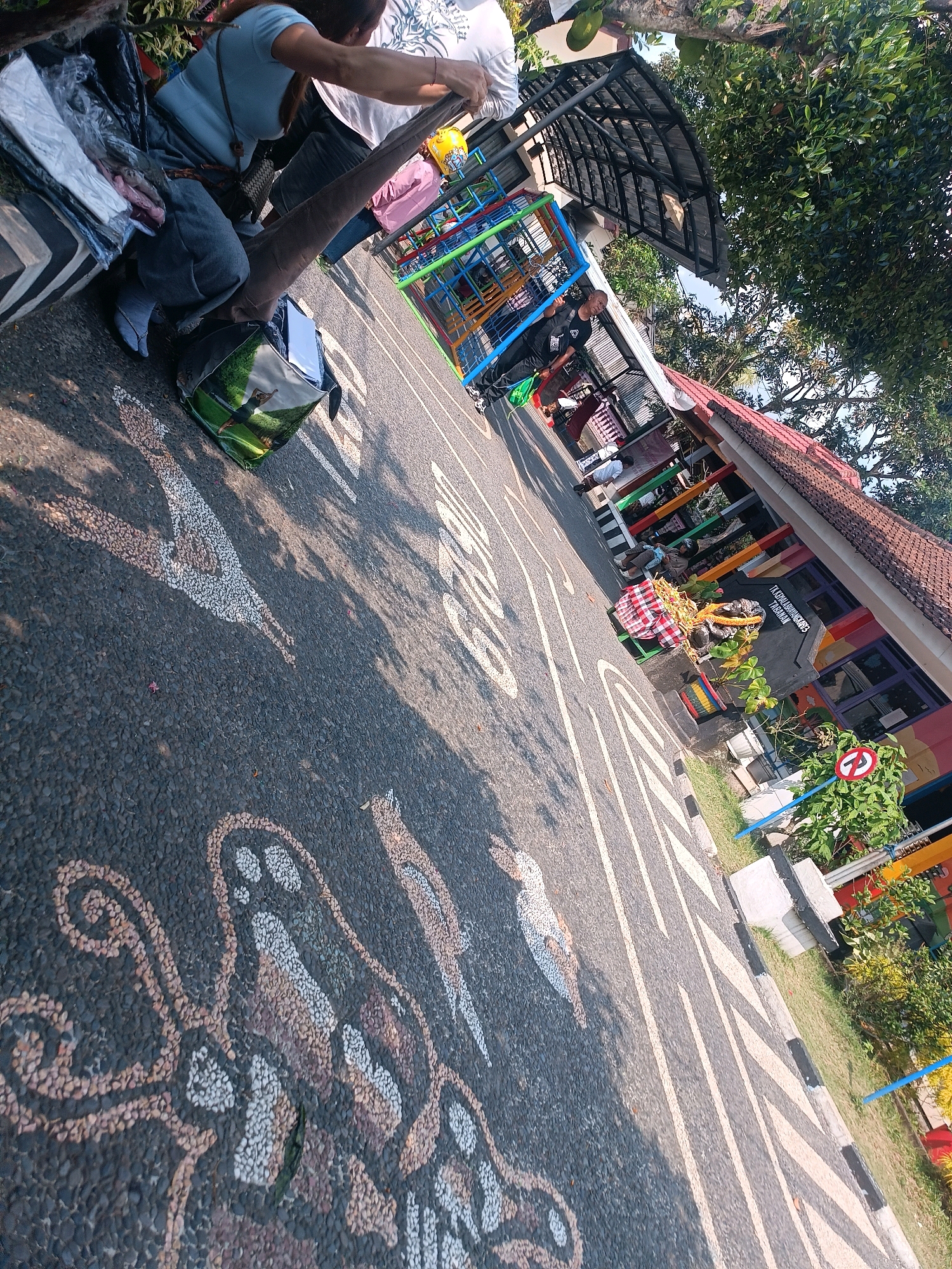 The image shows an outdoor scene on a sunny day, likely in Tabanan, Indonesia. In the foreground, the asphalt ground is decorated with white and brown pebble mosaics that appear to be stylized bird and floral motifs. The ground also has white painted lines, some of which are casting long shadows from overhead trees.
To the left of the frame, a person in a light blue t-shirt and grey pants is sitting on a bench with a black bag next to them. Another person in a white shirt is standing behind them, holding a long, dark brown fabric. A colorful child's playground structure is visible in the background, along with a building with a red tiled roof and colorful window frames. A sign on the building reads "TK KEPALA DESA MUNGGUHARJO TABANAN". A red and white checkered cloth is draped over a small green chair in front of the building, and a small traffic sign indicating "no right turn" is visible on the right side of the frame. The environment appears to be a public space, possibly a school or community center, with vegetation and trees surrounding the area.
