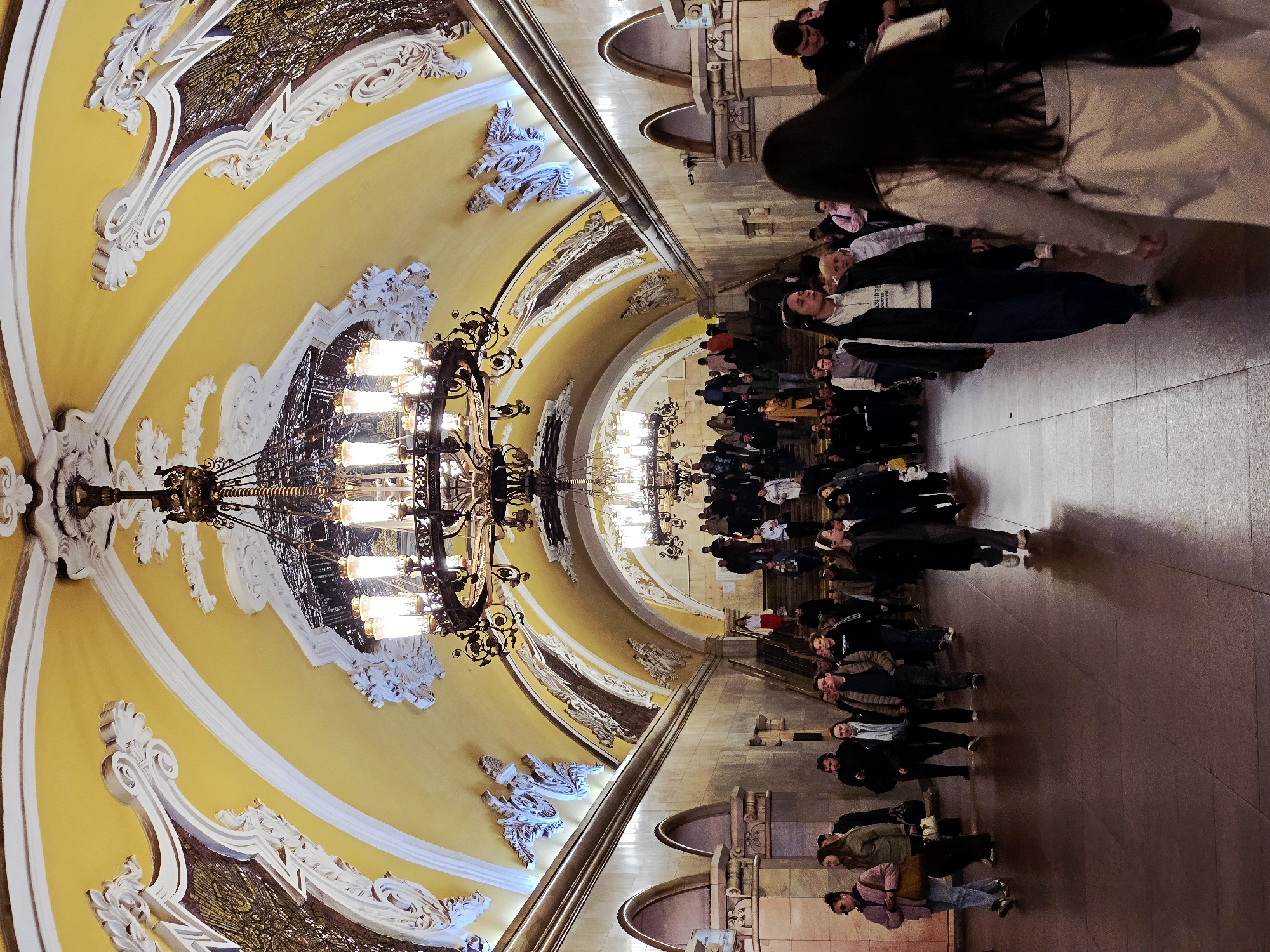 The image depicts the interior of the Kiyevskaya metro station in Moscow, Russia. The station features a vaulted ceiling painted in yellow and adorned with white decorative molding, including floral and geometric patterns. Large chandeliers hang from the ceiling.
A crowd of people, predominantly in dark clothing, is seen walking along the platform, which appears to be constructed from stone or tile. The platform is bordered by a railing with arched openings. The overall atmosphere suggests a busy, well-lit, and ornate public transportation space.