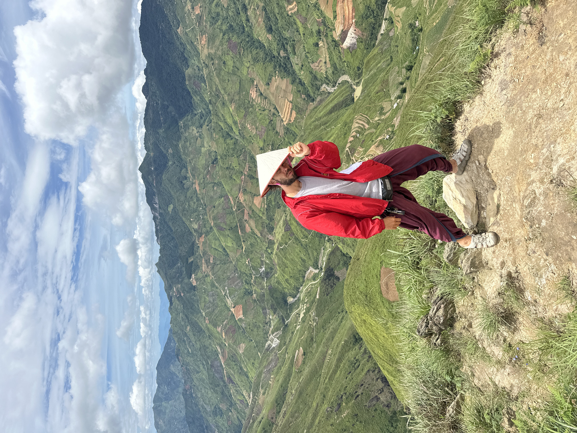 A man wearing a red jacket, maroon pants, and a conical hat stands on a rocky outcrop overlooking a vast, verdant valley in Ta Xua, Vietnam. The valley is characterized by a series of terraced hillsides, some cultivated and others covered in lush green vegetation, interspersed with winding paths and a visible river or stream. The sky is partly cloudy with a bright, diffused light. The man is posing for the camera, holding the brim of his hat with his right hand. The ground beneath him is a mixture of dirt, rocks, and sparse grass. The background showcases multiple layers of rolling mountains, fading into a hazy blue in the distance.