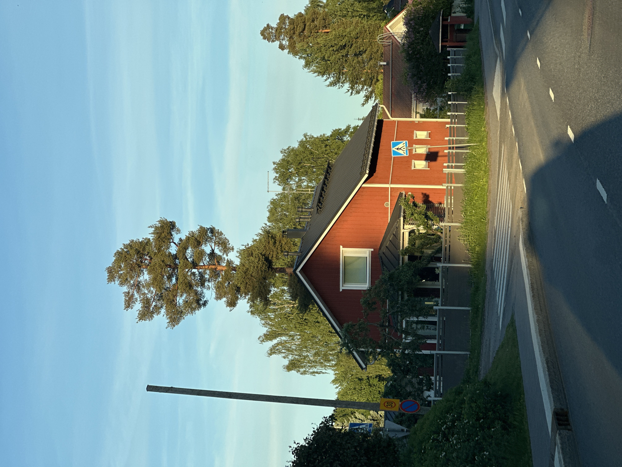 The image captures a quiet residential street scene bathed in the golden light of what appears to be late afternoon or early morning. A striking red wooden house with a black roof is the central focus. It features a single white-framed window on the visible side and a chimney stack. In front of the house, a grey wooden fence encloses a well-maintained garden area with lush green grass and trees. A pedestrian crossing is clearly marked on the asphalt road, with white stripes indicating the pathway.
Several road signs are visible: a blue and white pedestrian crossing sign, a circular red sign with a blue line, and a yellow sign with the number "30" inside a circle, likely indicating a speed limit. A tall, slender wooden pole stands prominently on the left side of the image, casting a long shadow.
Surrounding the house and fence are various trees of different heights and species, including a tall pine tree with a distinctive crown. In the background, other residential buildings and more trees are visible. The sky is a clear, vibrant blue with a few wispy clouds. The overall impression is one of peaceful suburban life in a well-kept environment.
