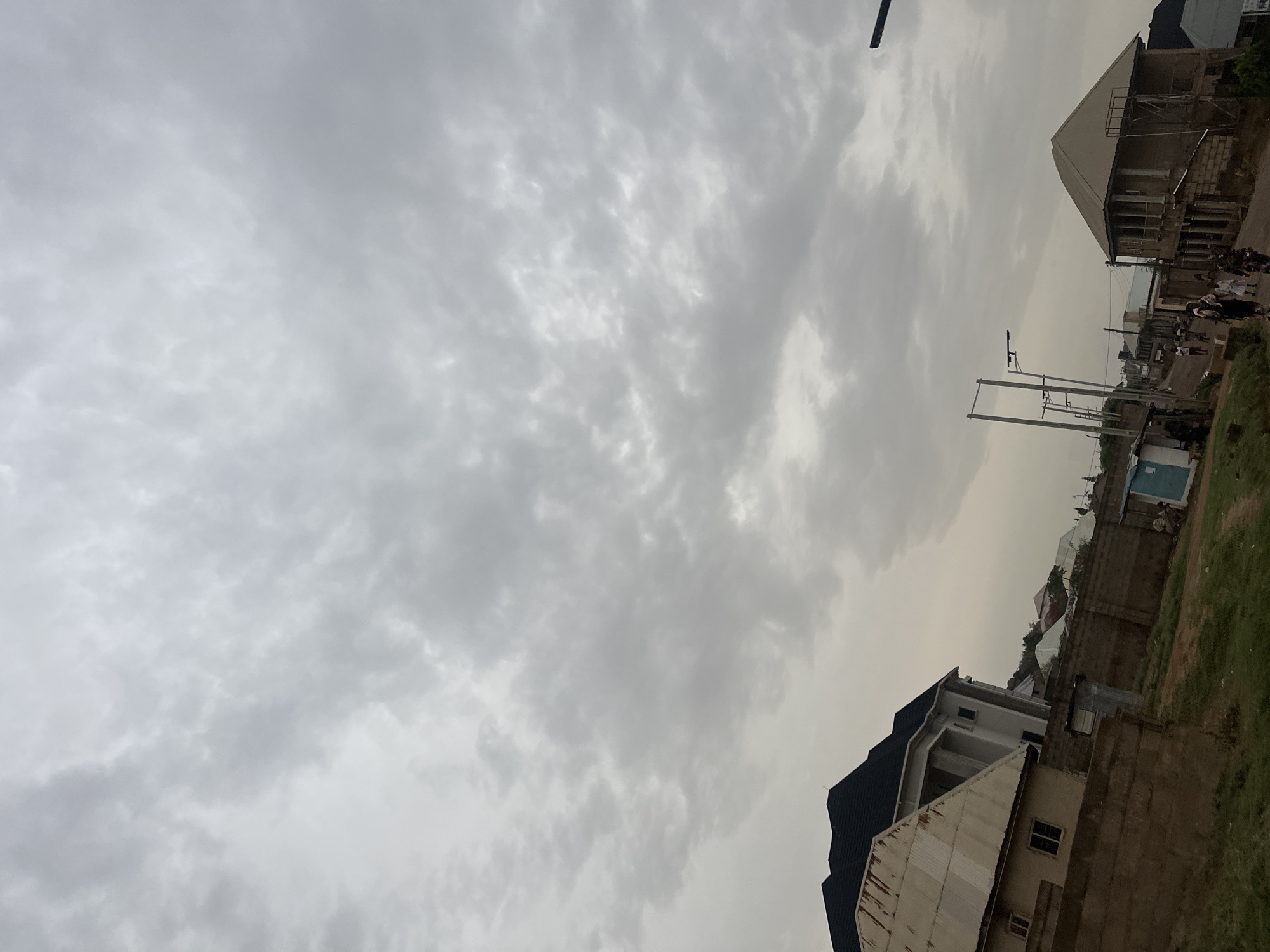 The image depicts an overcast sky above a street scene in Jos, Nigeria. The sky is filled with gray clouds. Buildings line the street, ranging from partially constructed structures to finished homes. A group of people is visible in the distance, appearing to walk along the street. Power lines are present, suspended by poles. The ground is partially covered with grass and foliage, suggesting a suburban or residential area. The image is rotated slightly, resulting in a skewed perspective.