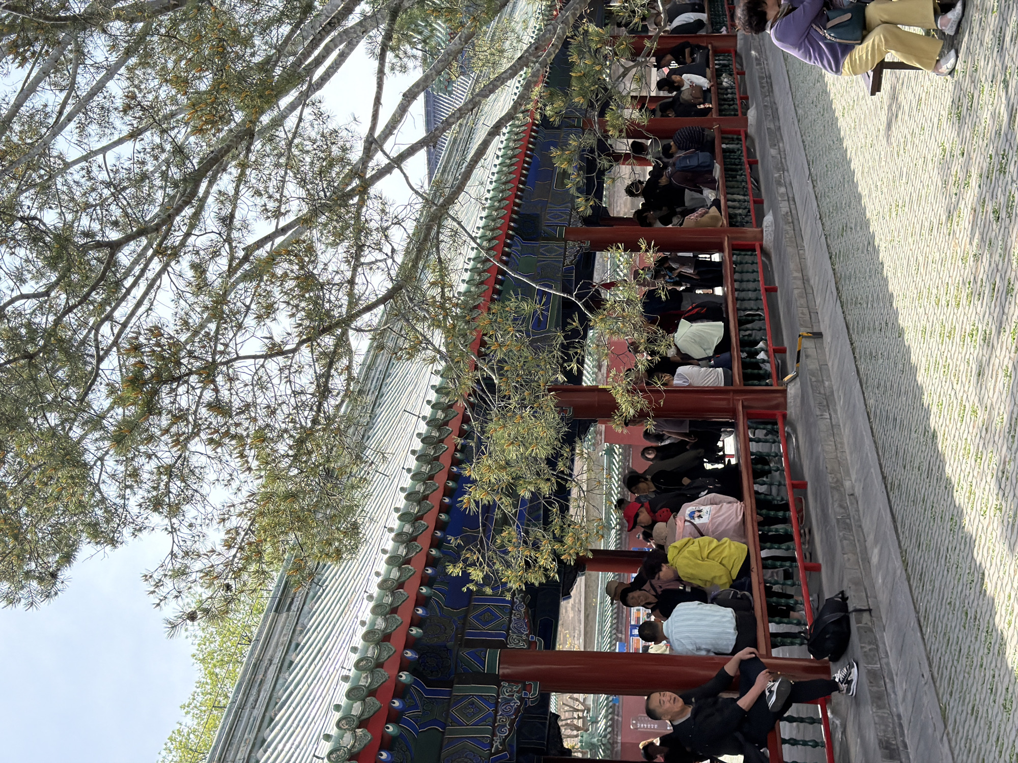The image shows an outdoor area in the Dongcheng District, China. The focus is a traditional Chinese structure with green-tiled roofing, ornate blue and red decorative elements, and a row of red wooden columns that create a covered walkway. The walkway is lined with people, mostly sitting, and some appear to be resting or observing. There is a gray paved ground to the right. Above the building is a tree with bare branches and budding leaves against a pale sky.