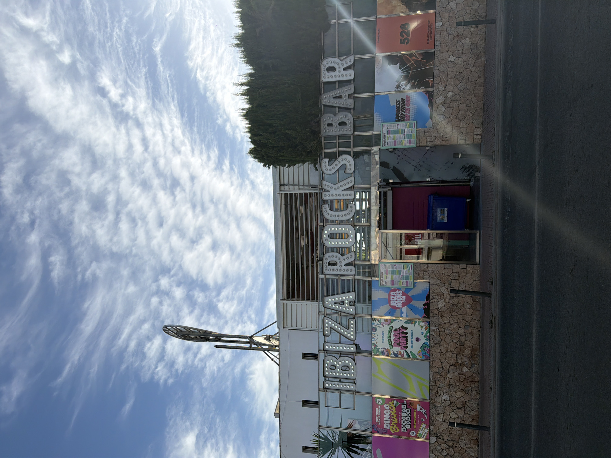 The image depicts the exterior of Ibiza Rocks Bar in Sant Antoni De Portmany, Spain, on a partly cloudy day. The prominent signage spells out "IBIZA ROCKS BAR" in large, illuminated letters. Below the main sign, a series of posters advertise events such as "Bingo Brunch," "Pool Party," and "Food Menu," alongside other promotional graphics. The building features a textured stone wall at its base and a dark asphalt road in the foreground. To the left of the entrance, a blue dumpster is visible. A tall, metallic structure with a circular element stands on the rooftop, and lush green trees are planted behind the building.
