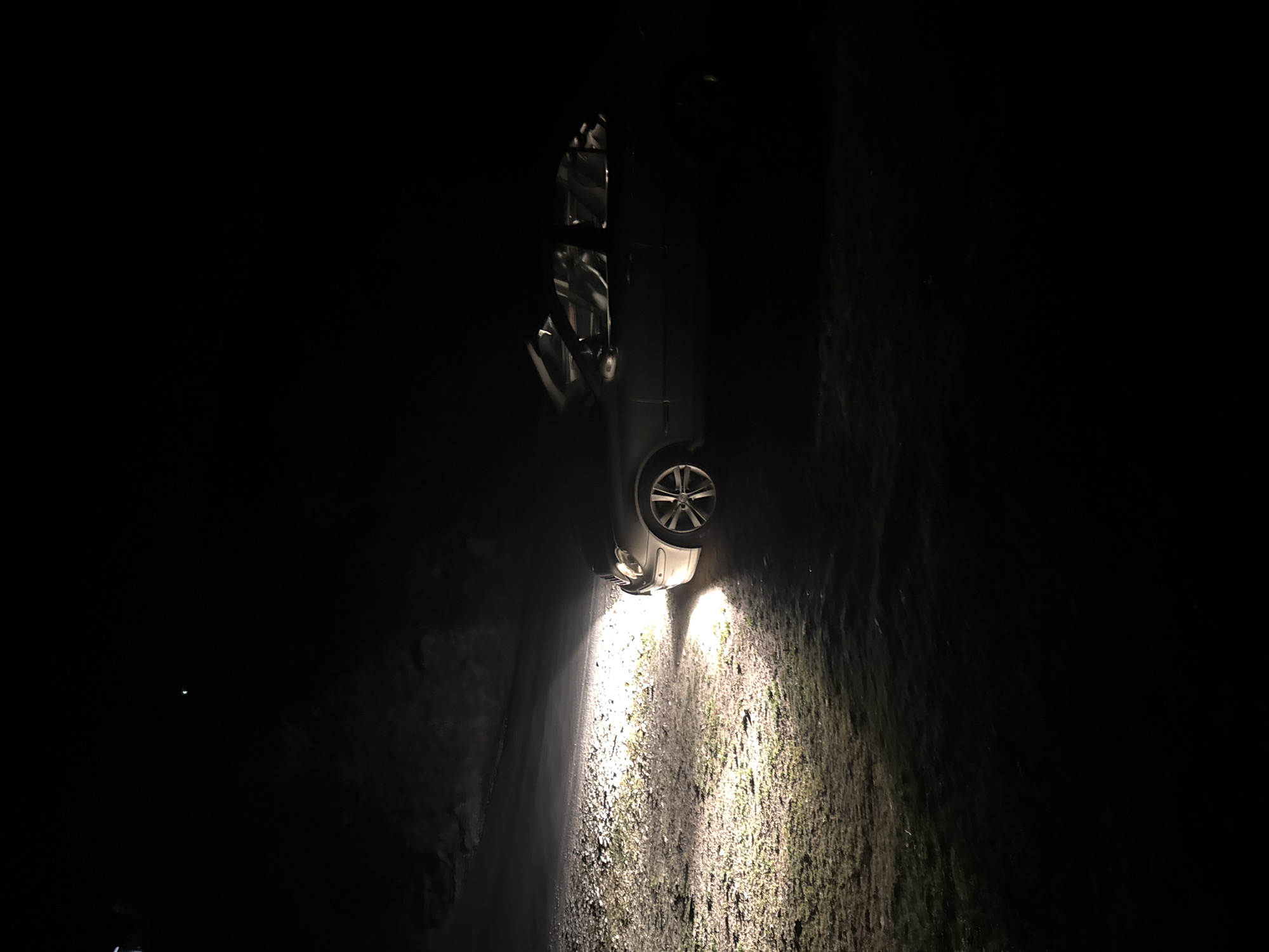 A car has crashed on a dark road, illuminating a mossy embankment in Sveti Konstantin, Bulgaria. The vehicle is upright, with its headlights on.