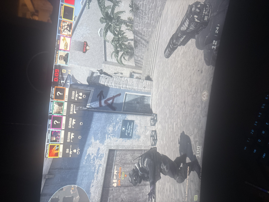 The image displays a computer monitor showing an active first-person shooter video game, observed to be played in the city of Yeroskipou, Cyprus. The in-game scene depicts an outdoor urban environment with light-colored stone buildings and cobblestone paving.
In the foreground, the player's right hand and a black and white weapon, identifiable as a Desert Eagle pistol, are visible. An ammunition counter "12 | 241" is displayed below the weapon. To the lower-left, a third-person perspective shows a character in dark tactical attire, wearing a gas mask and holding a rifle, moving in a crouched or low stance. Above this character, the in-game name "jok-rehim" and a money value "$100" are present.
The background features weathered stone architecture, including a building with a large red "A" and an arrow painted on its facade, indicating a game objective. Below this "A," a blue sign with Arabic text and "Biteb Ticket Sales" in English is affixed to the wall. Two dark-colored crates are positioned on the ground near the base of the building. To the right, a wide set of stone steps ascends, and several green palm trees are visible beyond the structures. A figure is seen clinging to the side of the building on the right.
The top section of the screen displays the game's user interface. A roster of player names includes "Coachen," "oskar," "MMB," "MI BOMBO," "jok-rehim," "Benjamin n.," "SSS," and "Saragosa." Health indicators all show "100." Monetary values for the first five players are "$0," "$2000," "$2050," "$3650," and "$100," respectively. A game timer reads "0:09," and the current score is "1 | 0." Additional interface elements include a prompt "100 in 3" on the right and "for 5 eliminated terrorists" text in the lower-left.
