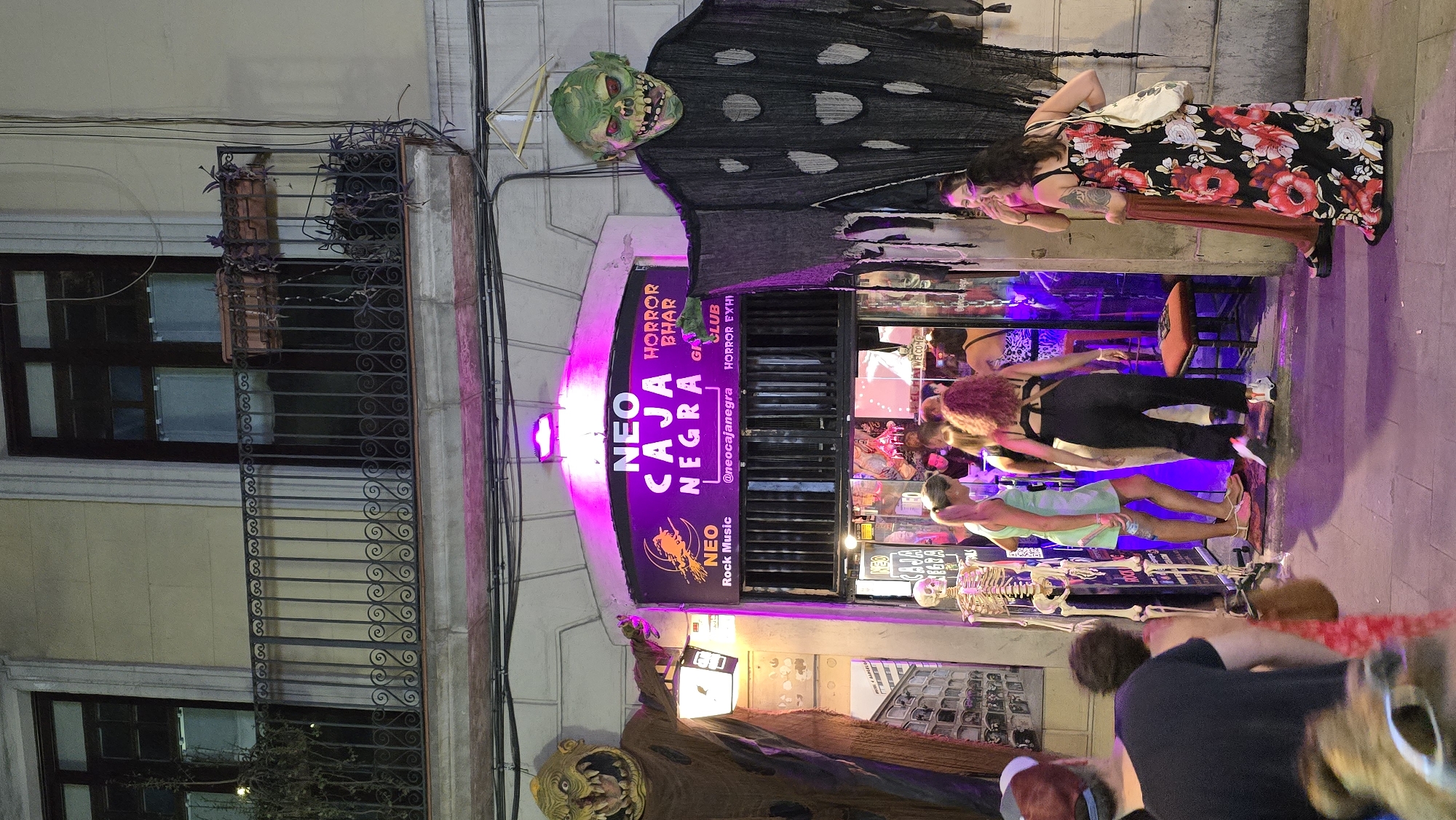 In Barcelona, Spain, in front of a nightclub called "Neo Caja Negra," a group of people are gathered. The nightclub has a Halloween theme, with two large monster decorations flanking the entrance. A skeleton stands near the doorway, and the interior is illuminated with purple lights. Several women are visible outside the club, some standing and talking while others appear to be entering or exiting. The overall atmosphere suggests a night out at a themed entertainment venue.
