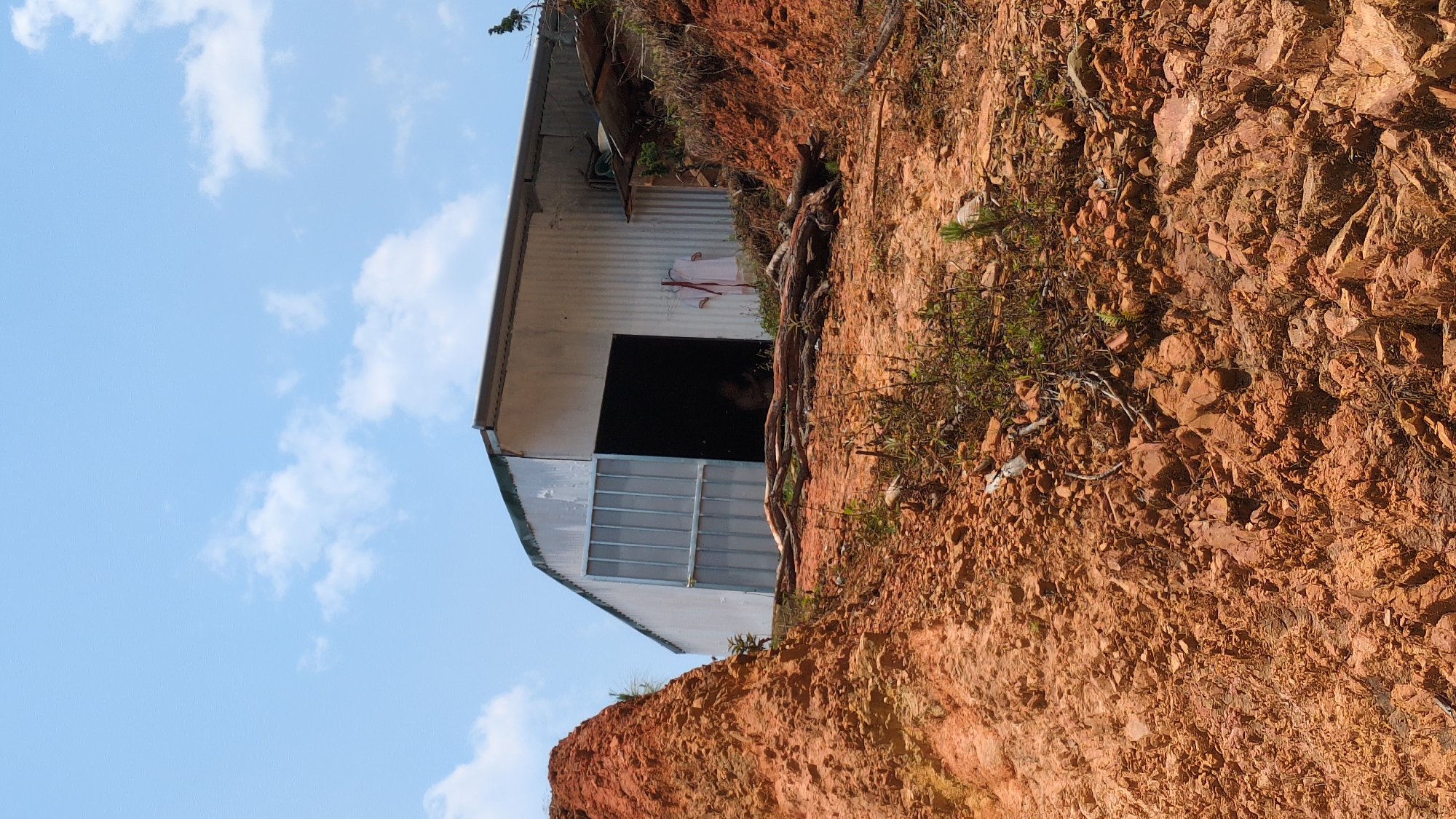 The image shows a shed-like structure built into the side of a red, rocky hill. The structure is made of corrugated metal, with a large, dark opening. The hill appears to be made of loose soil and rocks. The sky is blue with scattered clouds. The location is Lạc Dương, Vietnam. No people or interactions are visible.
