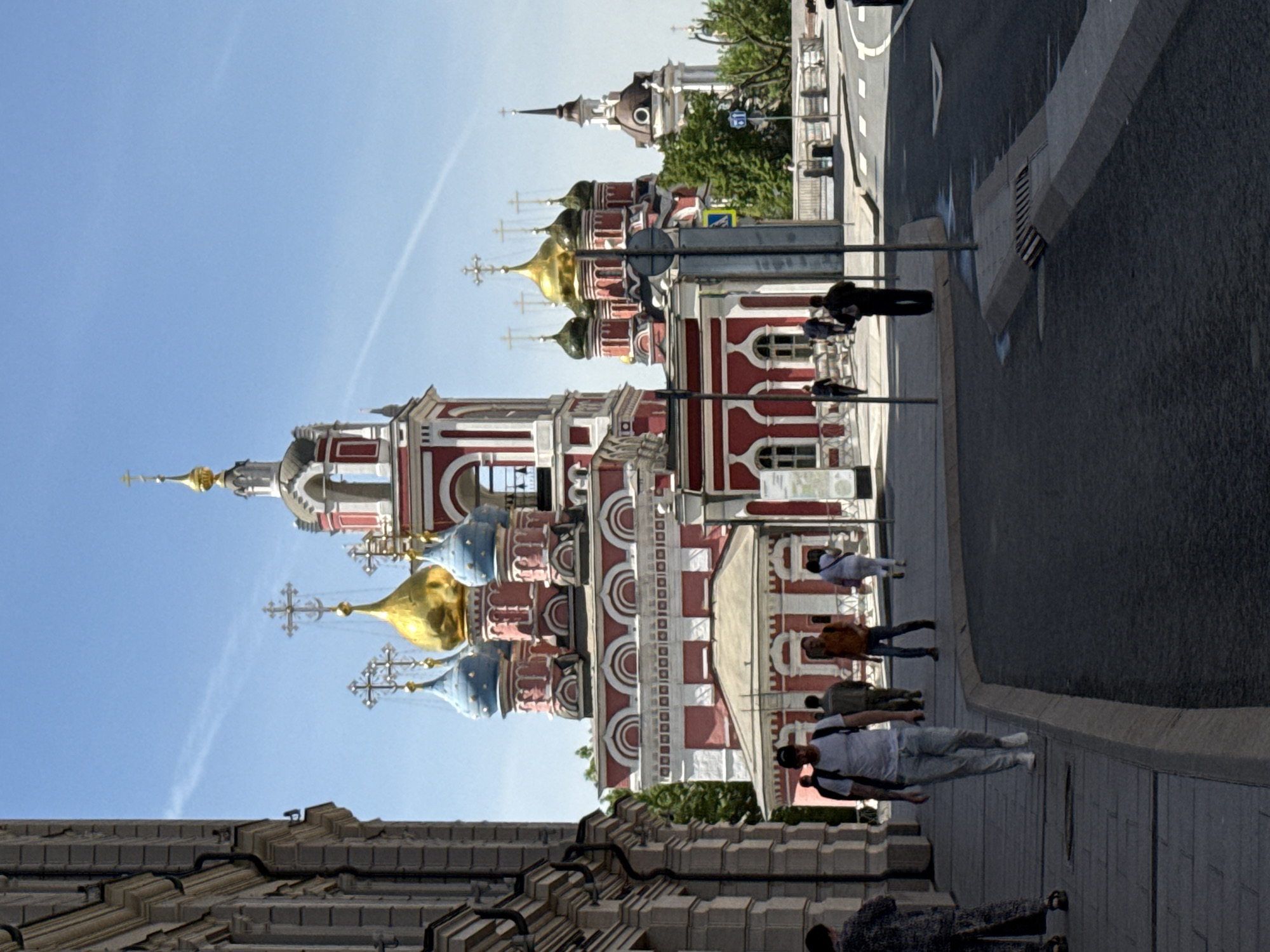 The image shows the Vysoko-Petrovsky Monastery in Moscow, Russia. The monastery complex is a prominent architectural landmark with colorful red and white facades, golden domes, and ornate crosses. People are walking on the sidewalk next to the building and along the street. The sky is clear and blue. The scene captures a typical daytime view of this historical site in an urban environment.