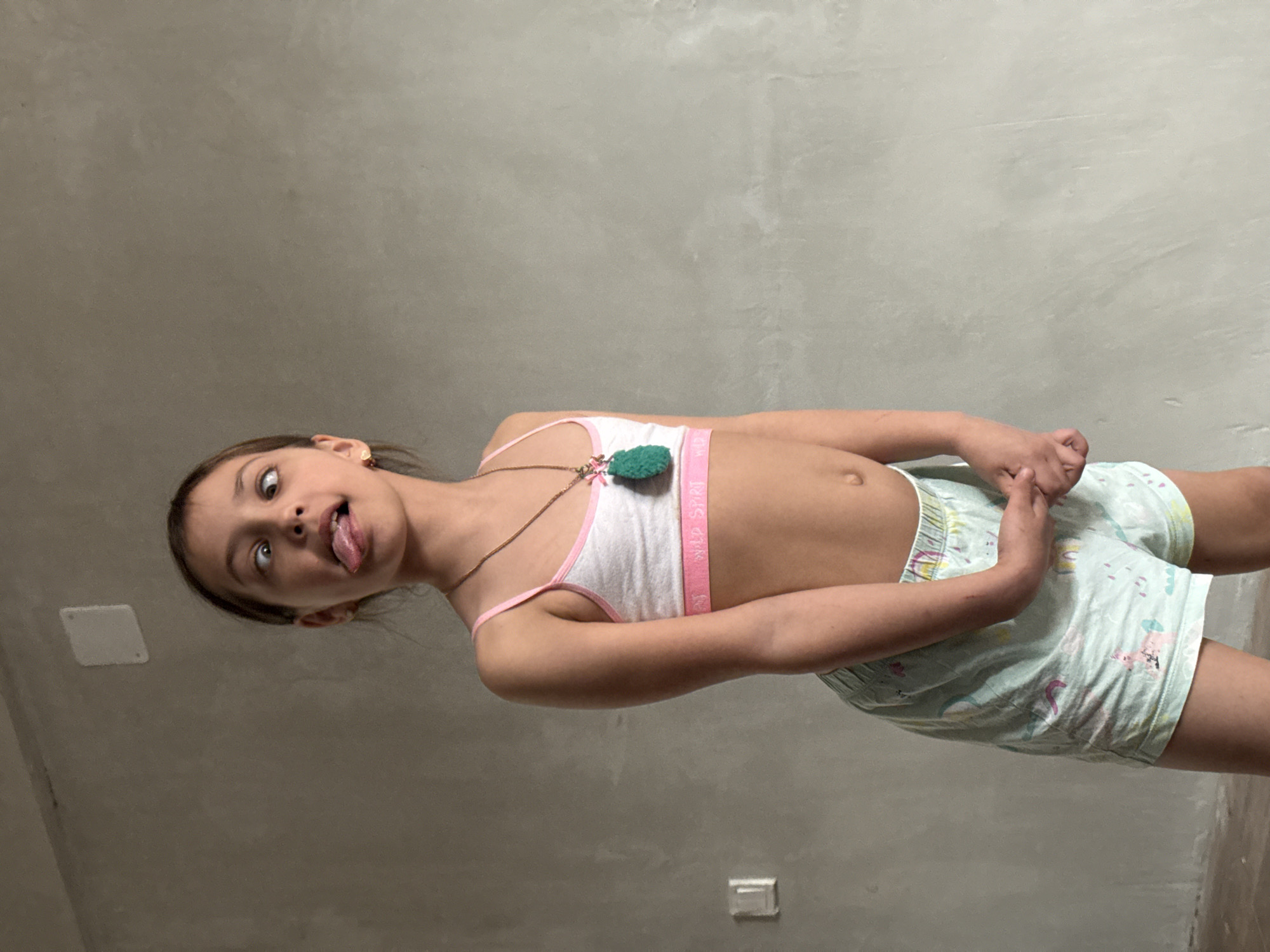 A young girl, approximately 7-10 years old, is the central subject of an indoor photograph, likely taken in Votkinsk, Russia. The image is oriented ninety degrees clockwise, depicting the girl striking a playful pose against a plain, light gray wall.
She looks directly at the camera with a mischievous expression, sticking out her tongue and looking slightly off-center with her eyes. Her light brown hair is pulled back. She is dressed in a white sports bra or crop top with pink trim; the elastic band across her chest displays partially visible text that appears to read "WILD SPORT." Around her neck, she wears a simple necklace with a distinctive green, fluffy, pompom-like pendant. She also wears light-colored shorts with faint, colorful patterns, possibly featuring cartoon characters or whimsical designs. Her hands are clasped together at waist level.
In the background, two square white objects, likely electrical outlets or light switches, are visible on the otherwise unadorned wall. The lighting suggests an indoor setting with artificial illumination. The overall mood conveyed by the girl's expression is one of lightheartedness and playfulness.