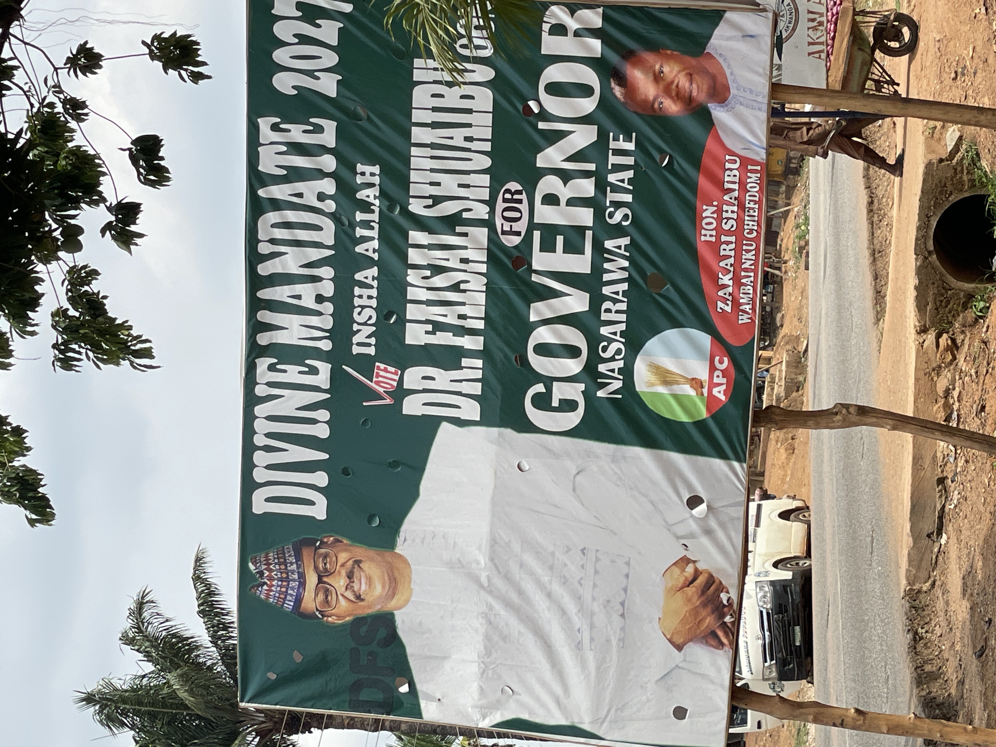 A large, weathered green political banner is displayed outdoors, likely alongside a road in Akwanga, Nigeria. The banner promotes Dr. Faisal Shuaibu for Governor of Nasarawa State in the "DIVINE MANDATE 2027" election. His large portrait, showing him smiling, wearing glasses and a traditional cap, occupies the lower-left portion of the banner. The text "VOTE INSHA ALLAH" is visible near his name.
A smaller portrait on the upper right features Hon. Zakari Shaibu, identified as "WAMBAI NKU CHIEFDOM I." The logo of the All Progressives Congress (APC), featuring a broom symbol, is positioned below his portrait. The banner is supported by wooden poles and shows signs of wear, including several small holes.
The background reveals a dirt road on the right, with a partially visible white van or bus and a concrete drainage pipe. An overhead overcast sky and lush tropical foliage, including a palm tree, frame the banner. A small cart or stall bearing the word "AKWANGA" is faintly visible behind the upper-right section of the banner.