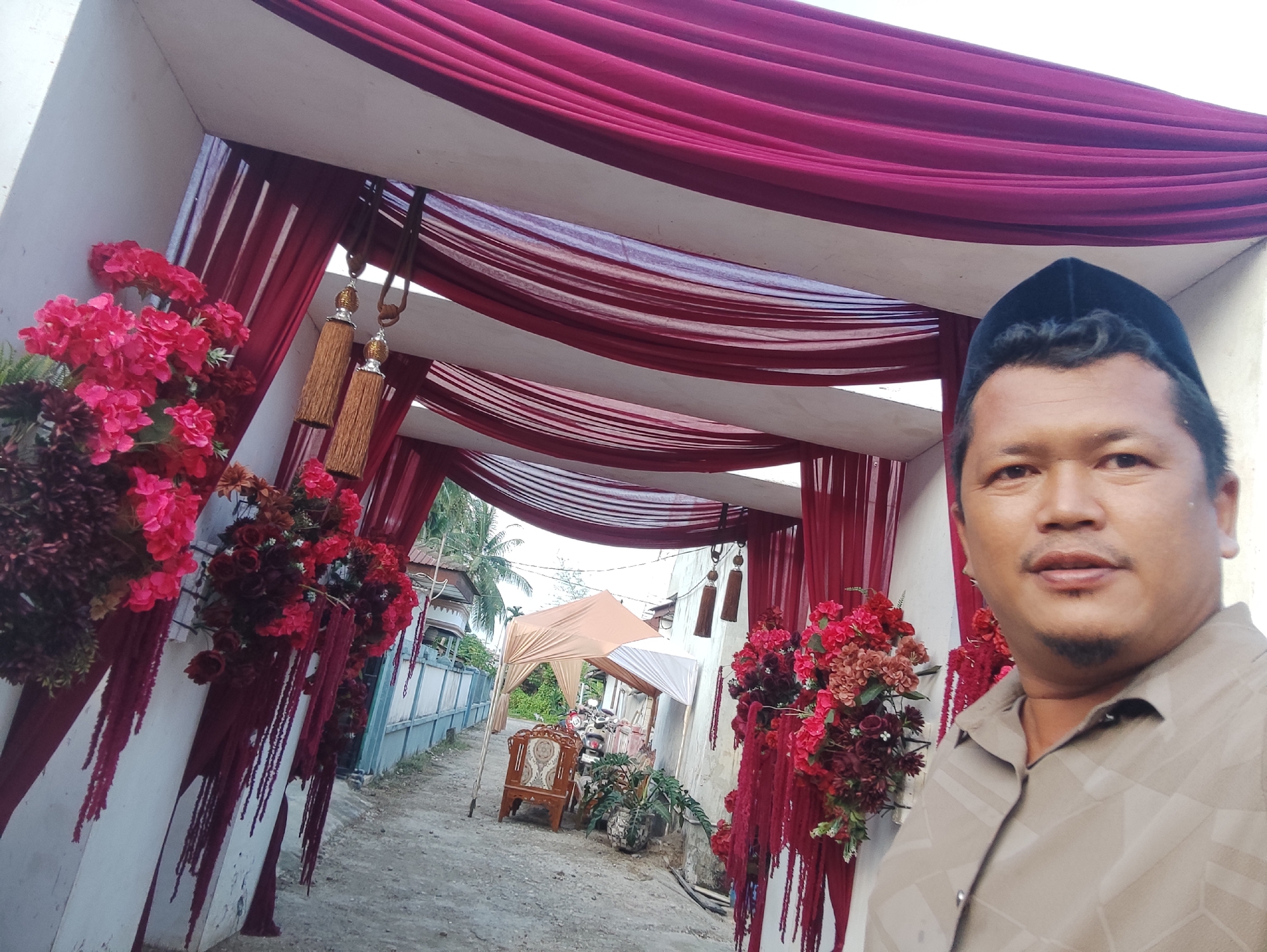 The image shows a man in a brown shirt and black cap standing in front of a decorated entrance, possibly for a wedding or celebration, in Gampongjawa, Indonesia. The entrance is adorned with red and pink fabric drapes, floral arrangements, and tassels. Beyond the entrance, a pathway leads to a tent-like structure with a motorcycle parked nearby. The scene suggests an event setting in a residential area.