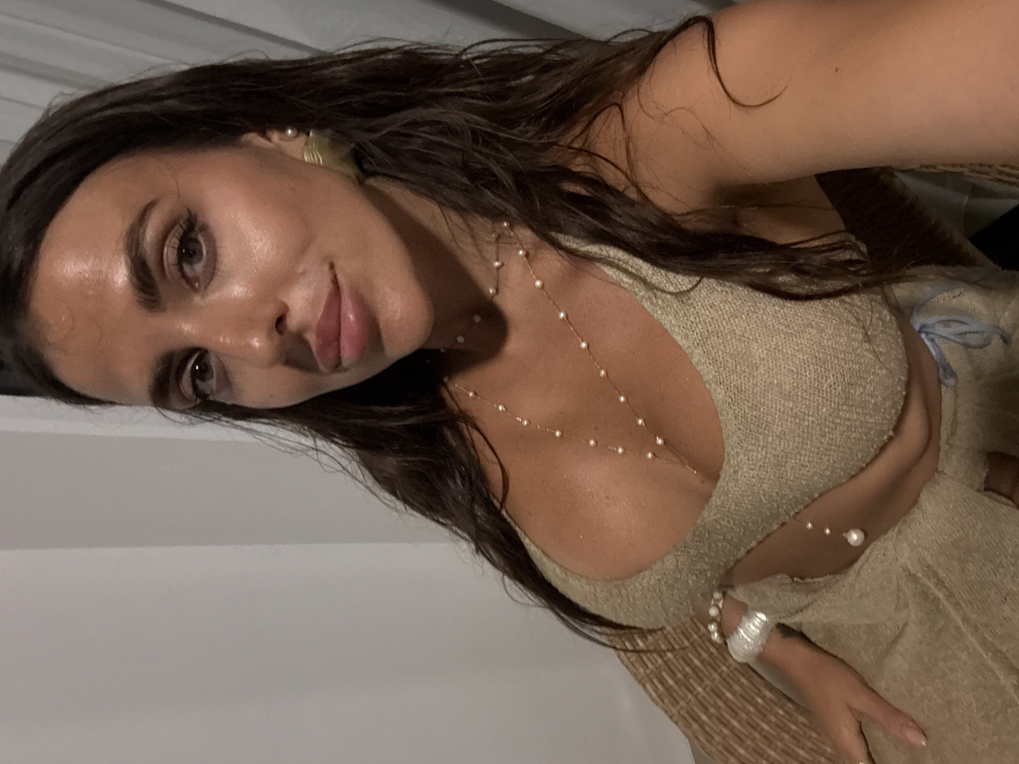 A tanned woman with dark hair poses indoors. She is wearing a beige textured garment with a plunging neckline, adorned with a pearl necklace and a bracelet on her wrist. The location is unspecified but presumed to be Kuta Selatan, Indonesia.