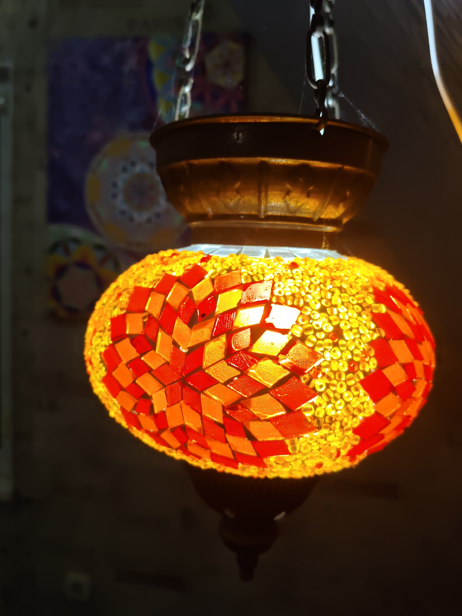 An ornate mosaic lamp hangs from a chain. The lamp's globe is covered in small, glass tiles in shades of red and orange, interspersed with tiny, yellow beads that glow warmly from the internal light. The mosaic pattern forms diamond shapes and starbursts. Above the globe, a darker, metallic fixture holds the chain. In the blurred background, what appears to be artwork or decorative panels with circular motifs are visible. The overall lighting is dim, with the lamp being the primary light source. There are no people or animals in the image. The setting is indoors, likely a shop or home, given the display of decorative items. The image is an artifact of urban life in Rostov-on-Don, Russia.