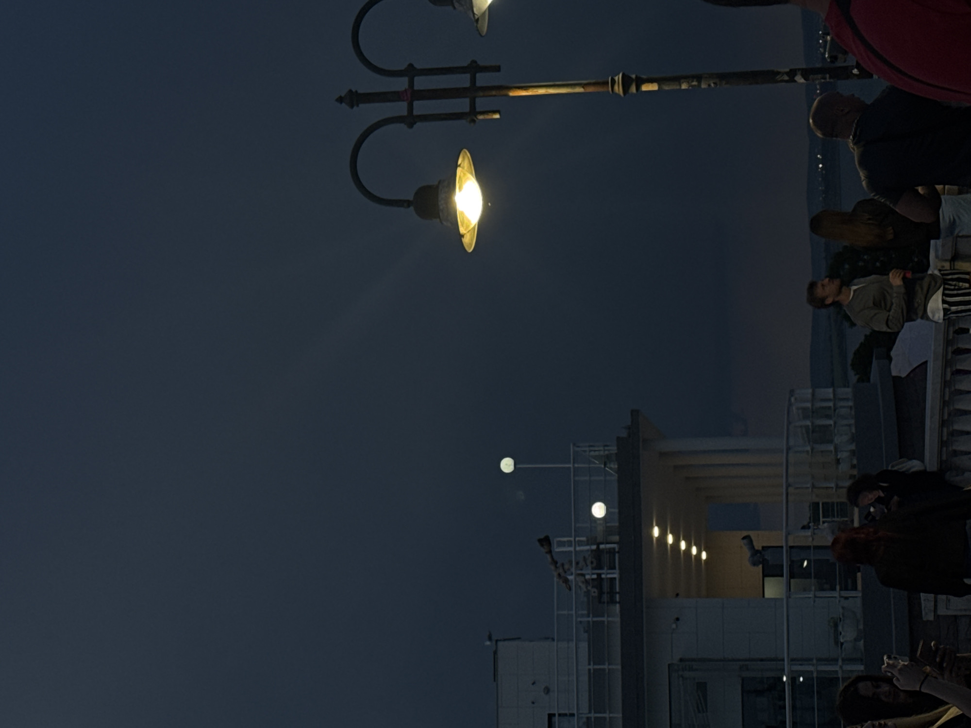The image depicts a nighttime scene in Burgas, Bulgaria. A vintage lamppost with two lit lamps is visible on the right side of the frame, positioned among a crowd of people. The crowd appears to be observing something, although the focal point of their attention is not clear in the image.
In the background, architecture features a modern building with illuminated lights along its facade. The scene is set against a dark sky. The overall ambiance suggests an evening gathering or event in an urban environment.