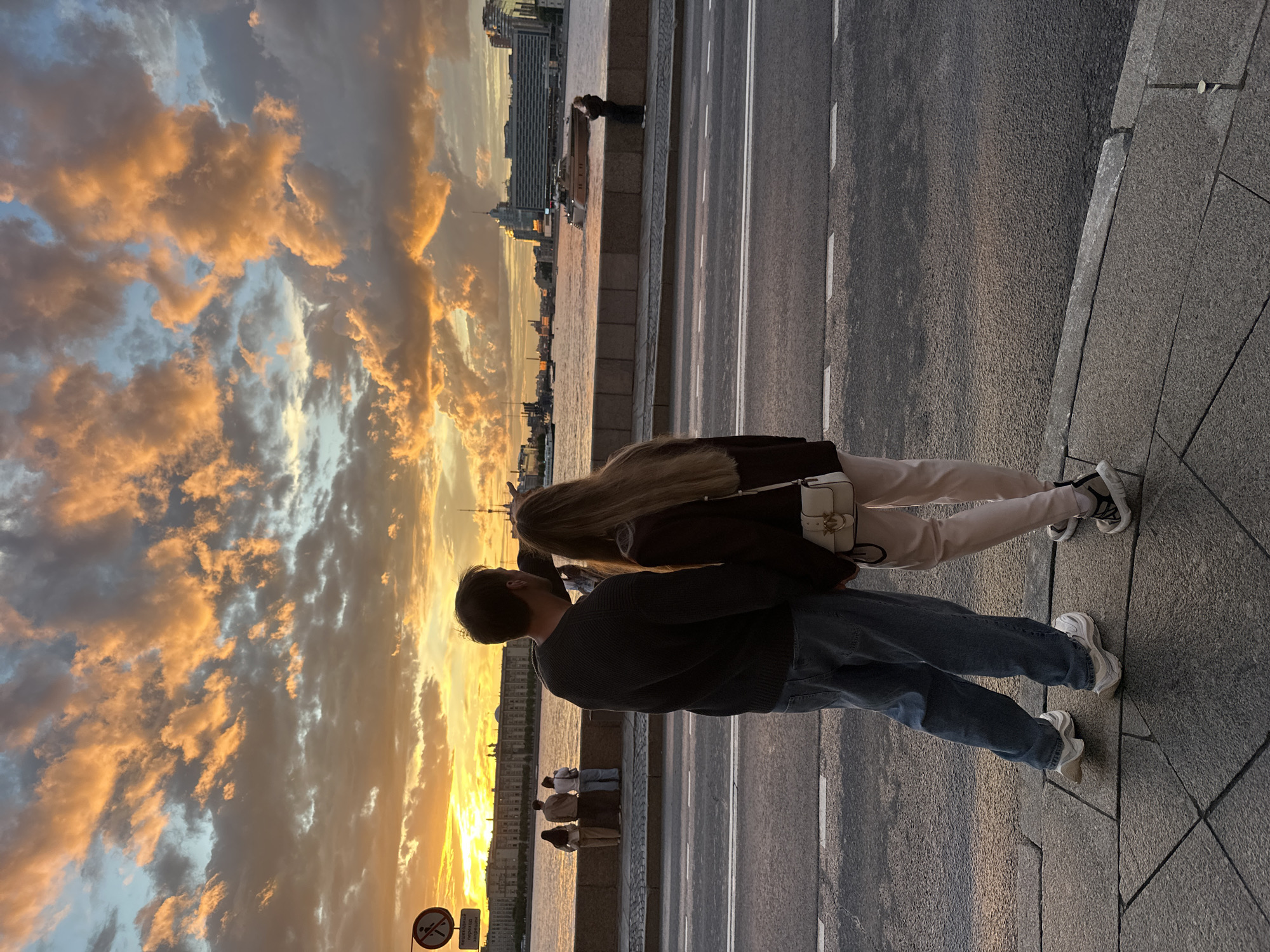 In Saint Petersburg, Russia, two individuals stand on a sidewalk overlooking a body of water at sunset. The sky is filled with dramatic, illuminated clouds in shades of orange and yellow, indicative of a beautiful evening. The cityscape of Saint Petersburg is visible in the background, with buildings lining the waterfront. The individuals are positioned facing away from the camera, looking towards the sunset. One person has long blonde hair, and the other has short dark hair. They appear to be casually dressed. The scene is peaceful, with a sense of shared appreciation for the natural spectacle. The lighting suggests it is late afternoon or early evening. There are no discernible emotions visible due to their posture, but the shared view of the sunset could imply a romantic or companionable moment. A sign with pictograms is visible in the foreground, but the text is not legible. The image captures a serene moment in an urban environment against a vibrant sunset sky.
