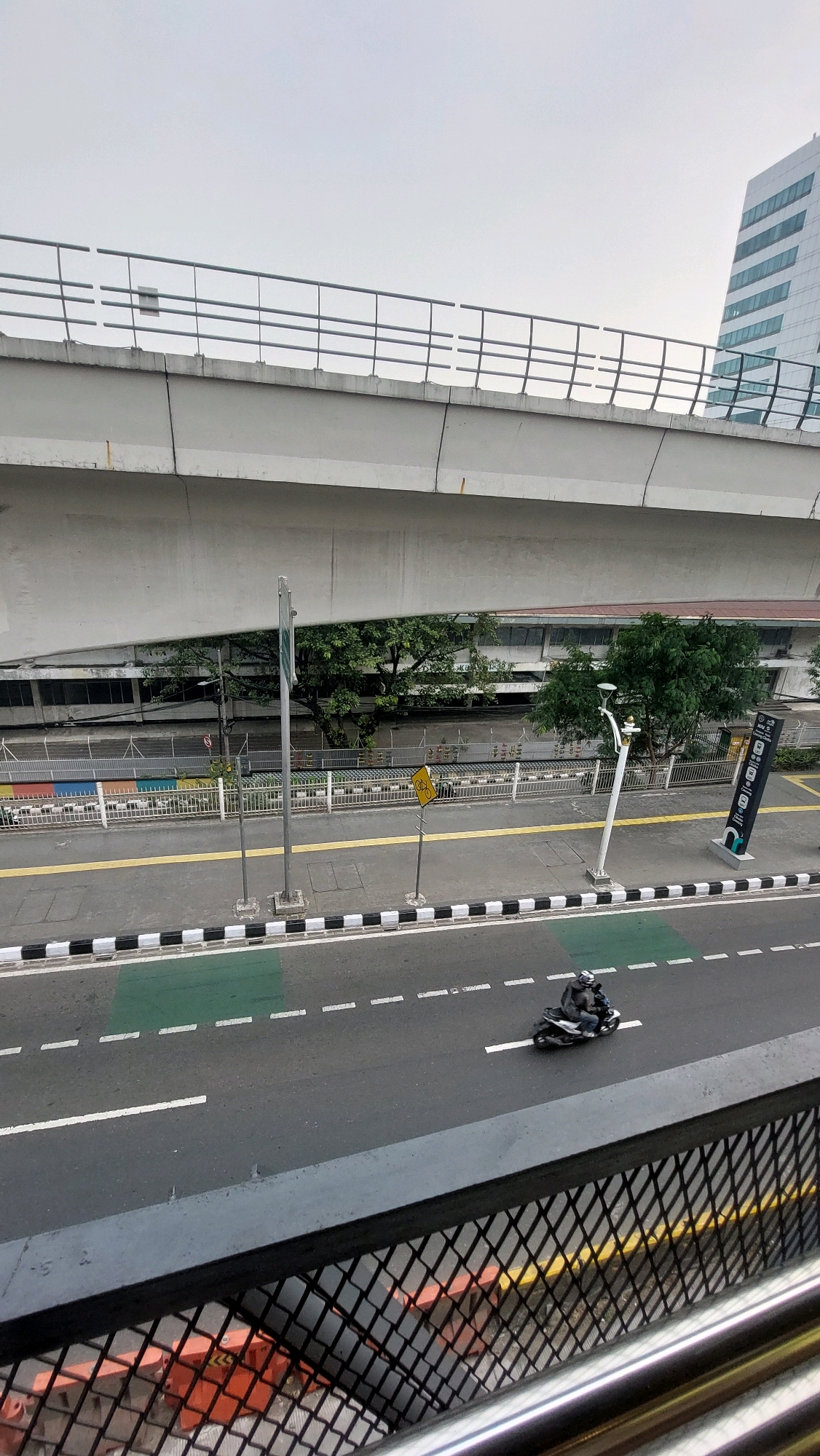 The image captures a view from an elevated walkway looking down onto a street and an overpass in South Jakarta, Indonesia. The scene is dominated by the grey concrete structure of the overpass, which spans across the top portion of the frame. Below the overpass, a multi-lane asphalt road is visible, marked with white dashed lines and a solid white line dividing the lanes. A portion of the road has a green painted section, likely a bike lane or a designated traffic area.
A single individual on a motorcycle is riding on the road, heading towards the right side of the frame. The motorcycle appears to be dark in color, and the rider is wearing dark clothing and a helmet.
To the left of the road, a sidewalk is present, demarcated by black and white striped curbing. A yellow line runs along the edge of the sidewalk. Several poles are present on the sidewalk, including a tall metal pole with a green sign and a shorter pole with a yellow traffic sign depicting a bicycle. Further down the sidewalk, another pole holds a directional sign with multiple icons.
Behind the sidewalk and a white metal fence, a row of trees with lush green foliage can be seen. Beyond the trees, a building with multiple windows is visible, appearing to be a commercial or residential structure. In the far right background, a tall, modern building with a white facade and angled windows stands out against the overcast sky.
The overall atmosphere suggests an urban environment on a cloudy day. The image is taken from a high vantage point, offering a broad perspective of the streetscape and its surrounding infrastructure.