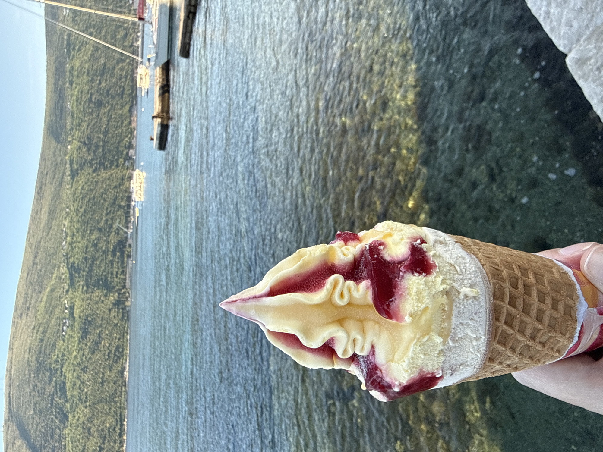 A close-up, slightly angled, outdoor shot shows an ice cream cone in the lower right foreground, being held in a hand. The ice cream is swirled, vanilla-colored, and drizzled with a reddish sauce. It sits atop a waffle cone. The background features water and a green, mountainous landscape under a clear sky, characteristic of a coastal setting. In the distance, there are boats and buildings along the shoreline, suggesting a marina or coastal town. The location is Đenovići, Montenegro.