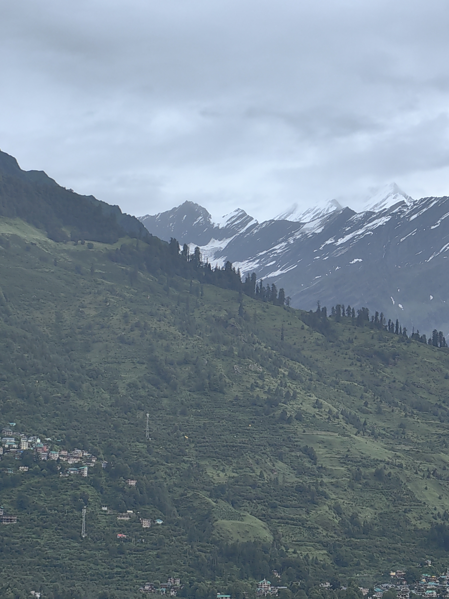 The image depicts a mountainous landscape with a village nestled on the slopes. The foreground features a steep, green mountain covered in trees and scattered houses. The houses are a mix of styles, with some appearing to be traditional and others more modern. Terraced fields are visible on the mountainside, indicating agriculture. In the midground, the mountain continues to rise, with more trees and buildings. The background is dominated by snow-capped mountains under a cloudy sky. The overall impression is one of a remote and natural setting. The location is Vashisht, India, a town known for its natural hot springs and proximity to the Himalayas.