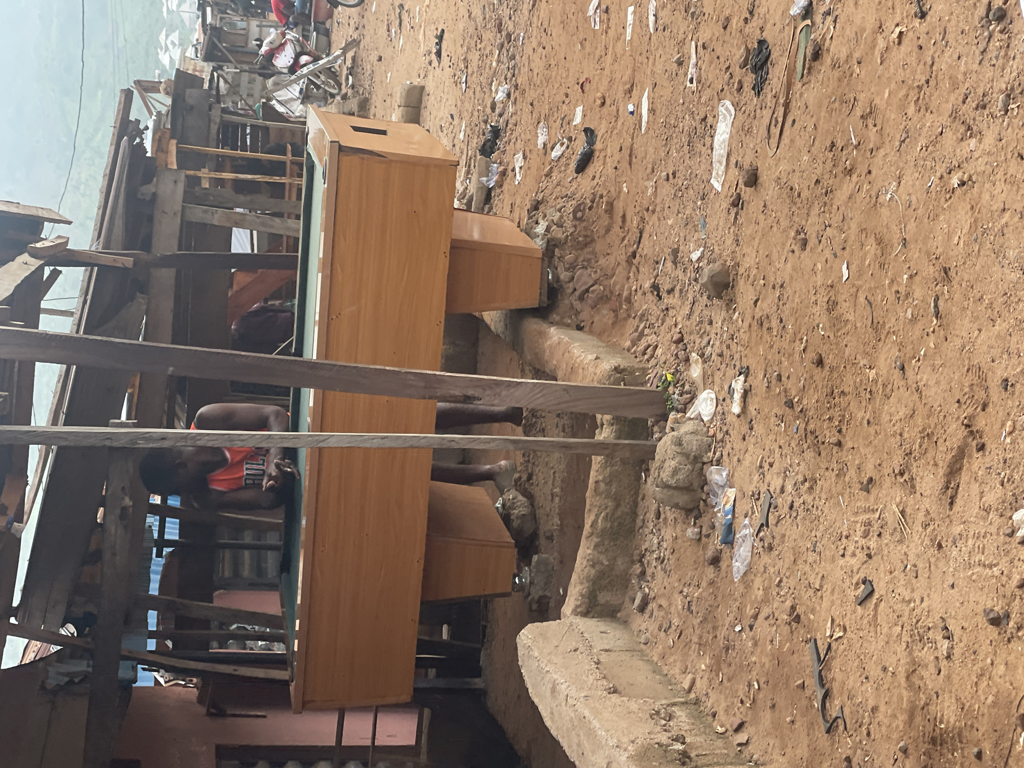 A person wearing an orange basketball jersey leans over a wooden billiard table situated on uneven, sandy ground. The billiard table has a green felt surface and is supported by four wooden legs. The surrounding environment appears to be an outdoor market or a makeshift structure, with rough wooden beams and corrugated metal roofing visible in the background. Debris, including plastic bags and scattered trash, litters the ground. A motorcycle is parked to the right of the scene. The background shows a hilly landscape with some structures on the slopes, consistent with the topography of Lokoja, Nigeria. The overall scene depicts a casual, possibly recreational setting within a somewhat unkempt urban or semi-urban environment.