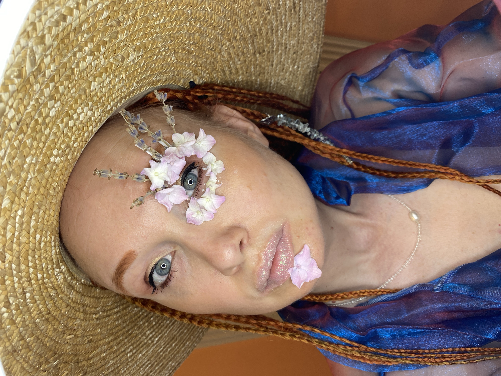 The image shows a close-up of a woman wearing a straw hat, with her face angled towards the camera. She has red hair in braids and is adorned with small pink and white flowers and lavender sprigs around her right eye and on her lower lip. She is wearing a sheer blue garment. The background is a solid orange color. The image appears to be taken indoors, with artificial lighting. There is no indication of the city of Sochi, Russia, in the image or its description.