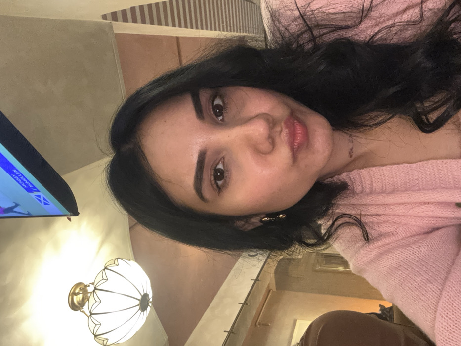 An adult female is depicted in a close-up, selfie-style photograph, taken indoors within the city of Howrah, India. She has long, dark, wavy hair, parted slightly off-center, and brown eyes. Her eyebrows are dark and well-defined. Her lips are pursed, forming a 'kissing' expression. She wears a light pink, knitted cardigan over a lighter-colored garment. A gold-toned dangle earring is visible on her left ear.

The background includes a decorative ceiling-mounted light fixture with a brass base and a white, fluted glass shade in the upper left. A wall-mounted television screen, displaying text including "MUNSEY," "MULLEN," and "SCOTLAND" with a flag graphic, is visible in the upper right. The ceiling is light-colored, and the walls exhibit light brown or beige tones with some vertical striped patterns. A dark, possibly upholstered element is partially visible on the far left. The lighting in the room appears warm. The subject is looking directly into the camera.