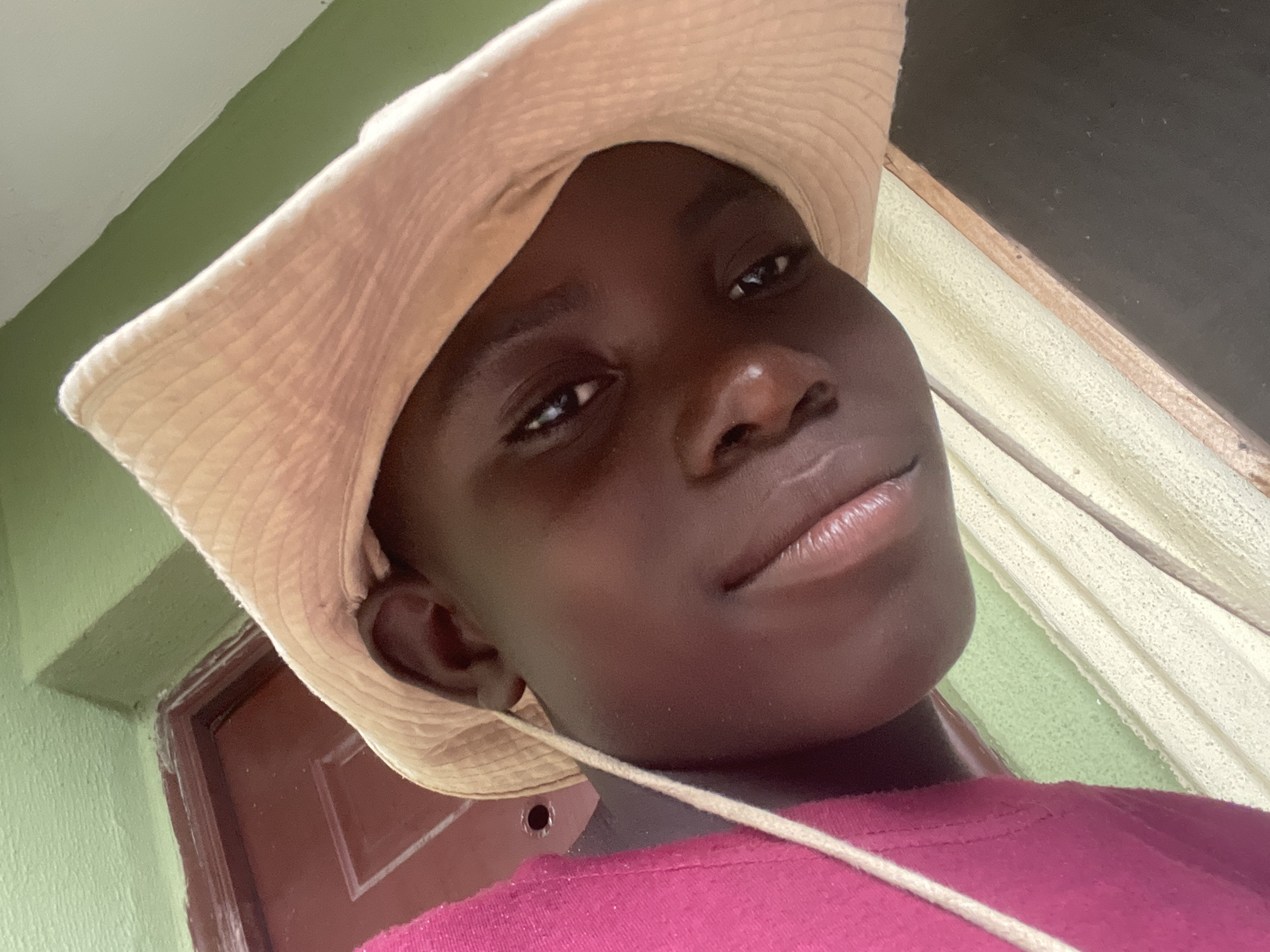 A close-up, low-angle selfie shows a young person with dark skin wearing a light-colored, straw-like hat and a bright pink shirt. The person's face is turned towards the camera, with their lips slightly parted. A thin, light-colored string hangs from the hat, crossing the person's chest. In the background, a red door with a white frame is visible on the left, and a textured, light green wall is above it. To the right of the person, a portion of a wooden structure or wall with a light-colored, textured surface is present, with a dark surface below it. The overall lighting suggests an outdoor or well-lit indoor environment. The scene appears to be a casual, personal photograph. The presence of the red door and green wall, as well as the wooden architectural elements, are consistent with residential or commercial structures found in Ado, Nigeria.