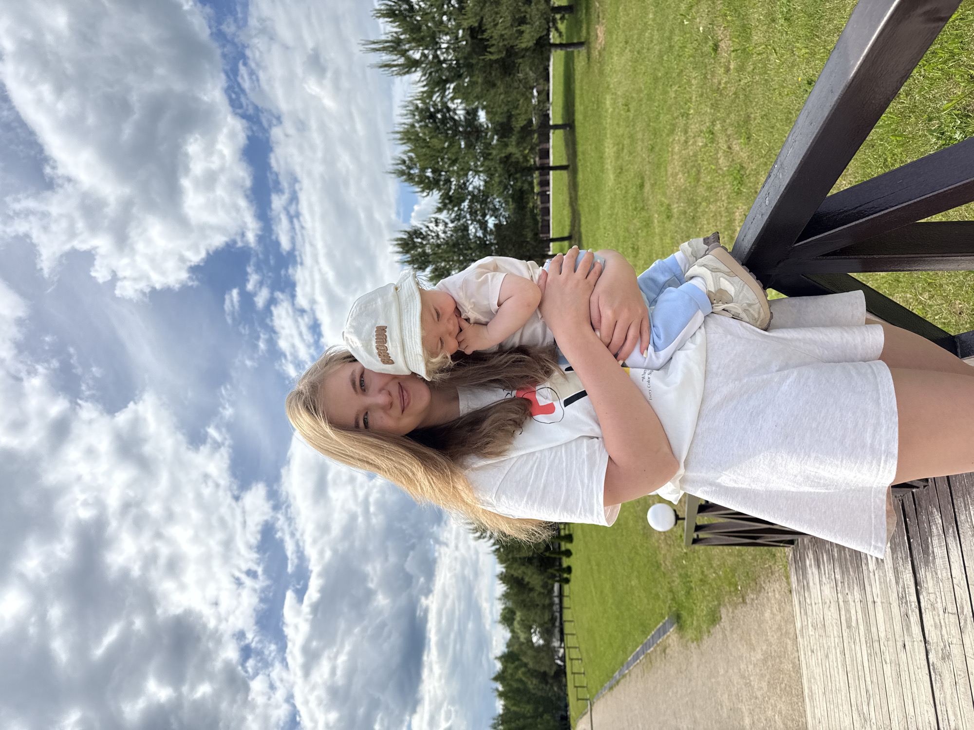 This brightly lit outdoor photograph, taken in Dobrograd, Russia, captures a young woman tenderly holding a baby. The image is presented sideways, suggesting it was captured in portrait orientation and then rotated.
The woman, with long, straight blonde hair, smiles gently as she looks towards the viewer. She wears a white short-sleeved t-shirt with a faint graphic design on the front and white shorts or a skirt. Cradled in her arms is an infant, dressed in a light-colored top, light blue bottoms, and light-colored sneakers with a thick sole. The baby wears a cream-colored bucket hat with the word "BROWN" clearly visible on the front. Its head rests against the woman's shoulder or chest, and its left arm is raised, partially obscuring its face, giving the impression of peaceful rest or sleep.
The outdoor setting features a vibrant backdrop. The sky above is a mix of bright blue with large, fluffy white clouds. A line of green trees marks the transition to a wide expanse of neatly mown green grass in the midground. To the right, a dark, sturdy railing or fence is prominent, suggesting the subjects are situated on an elevated wooden deck or pathway, parts of which are visible. A white spherical lamp post can also be seen near the railing.
The scene conveys a serene and loving moment between the woman and the baby, set on a pleasant, cloudy yet bright daytime.