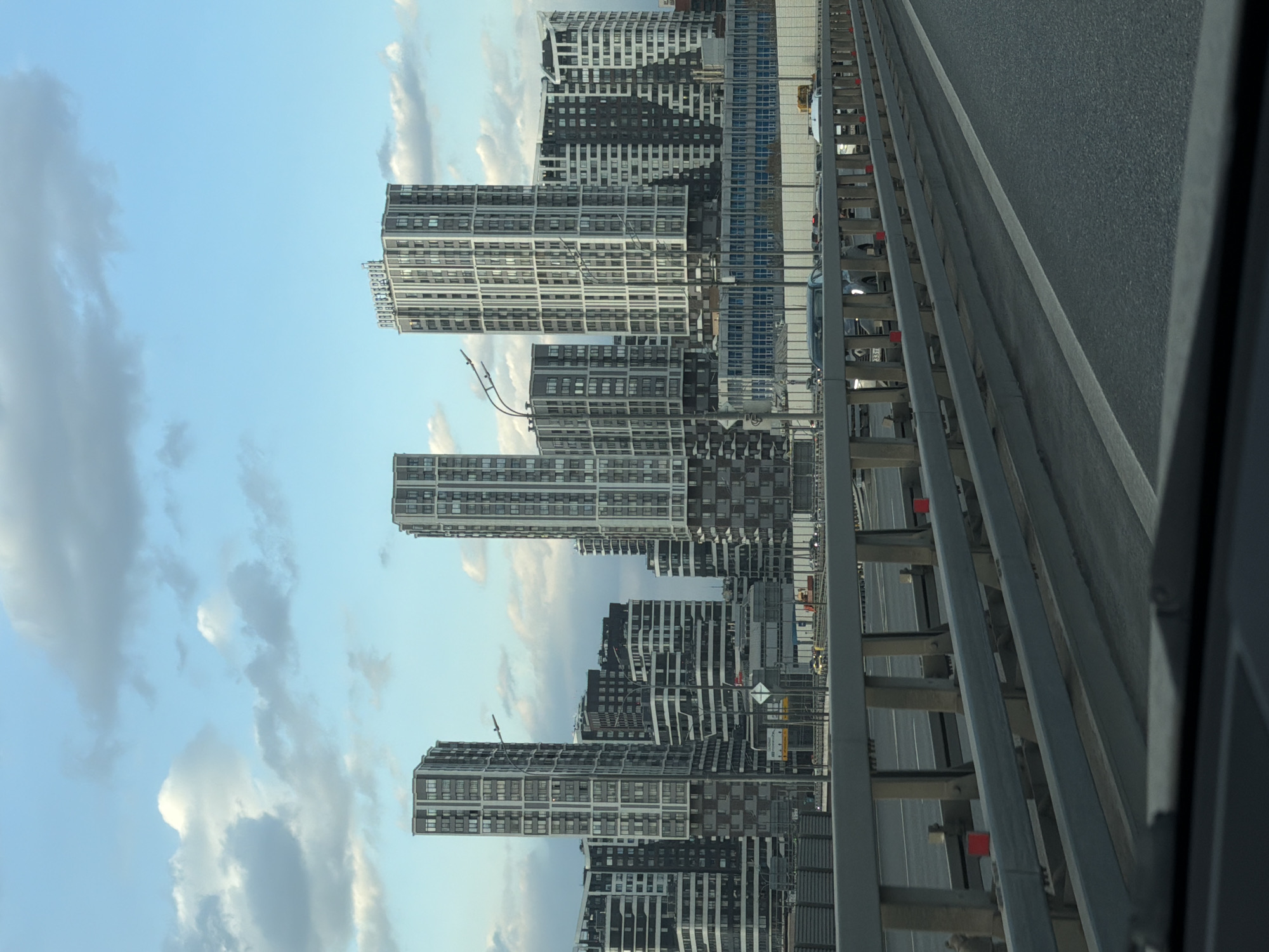 The image depicts a cityscape of Moscow, Russia, featuring modern high-rise residential buildings under a cloudy blue sky. The buildings are characterized by their geometric patterns and gray and white color schemes. A highway overpass is visible in the foreground, with vehicular traffic passing by. The photograph appears to have been taken from within a vehicle on the overpass.