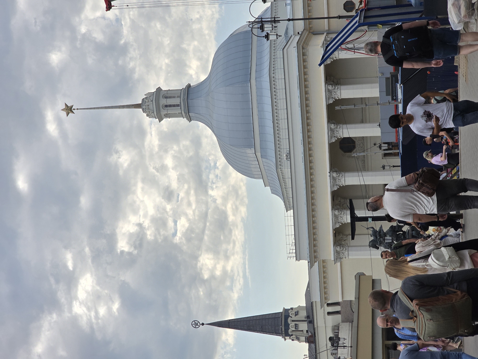 The image depicts a street scene in Moscow, Russia. Notable architectural features include a dome-shaped building with a gold star atop a spire, visible against a cloudy sky. People are seen walking along the sidewalk in front of a building with columns, suggesting a public or commercial area. Individuals are dressed in casual attire, indicative of a daytime setting. There are details such as flags, street lamps, and architectural ornamentation that provide further context about the location. The scene suggests a blend of tourism and everyday life in a metropolitan environment.