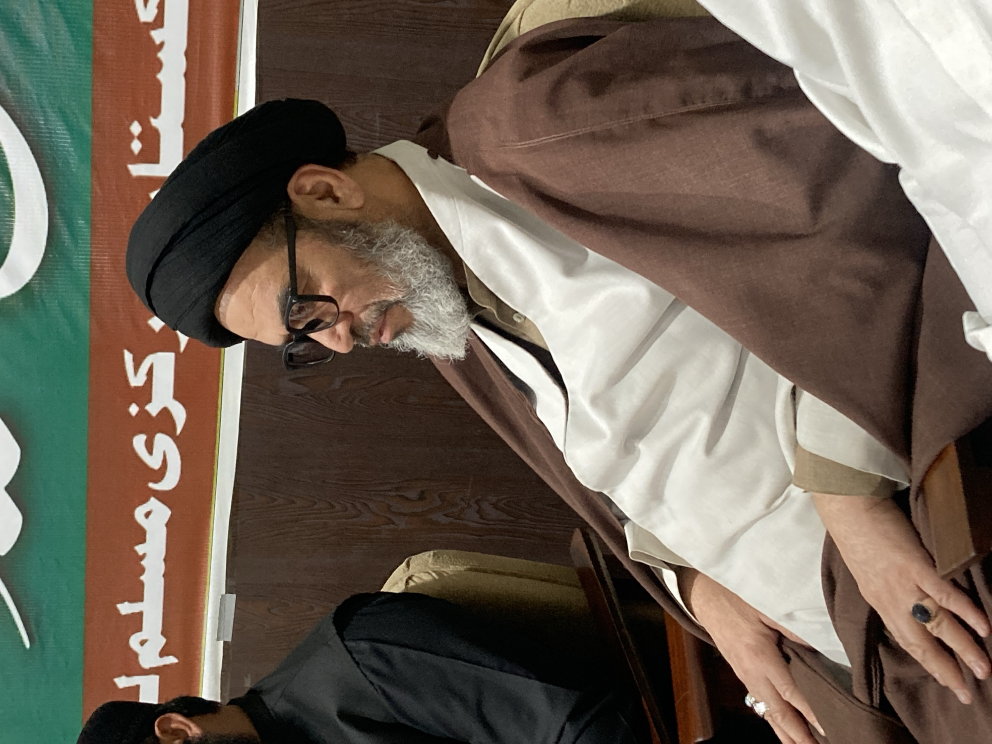 An elderly man with a white beard and black turban sits in a chair wearing glasses, a white shirt, and a brown robe. He has a ring on his right hand. Part of a green and white banner is visible on the left side of the image. The man is inside, possibly in Quetta City Tehsil, Pakistan.