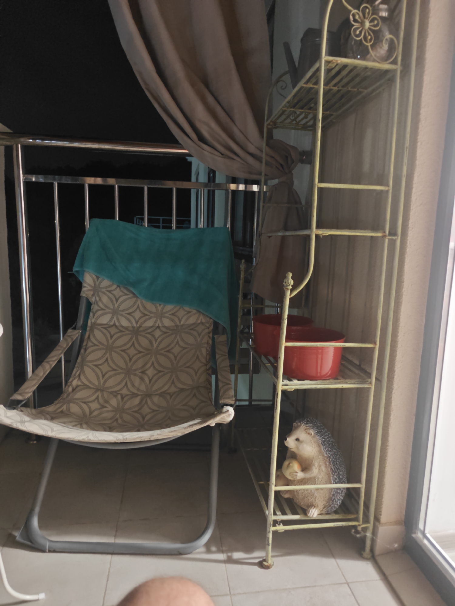 The image depicts an outdoor balcony at night. A beige and grey patterned folding chair is positioned prominently on the left, draped with a teal towel. To the right, a tall, pale yellow metal shelving unit stands against a textured wall. The shelves hold two red bowls on the second shelf from the bottom, and on the bottom shelf, a ceramic figurine of a hedgehog is holding a small yellow object. A metal railing runs along the edge of the balcony, with a dark curtain obscuring the background. The floor is tiled. The location is identified as Sukko, Russia.